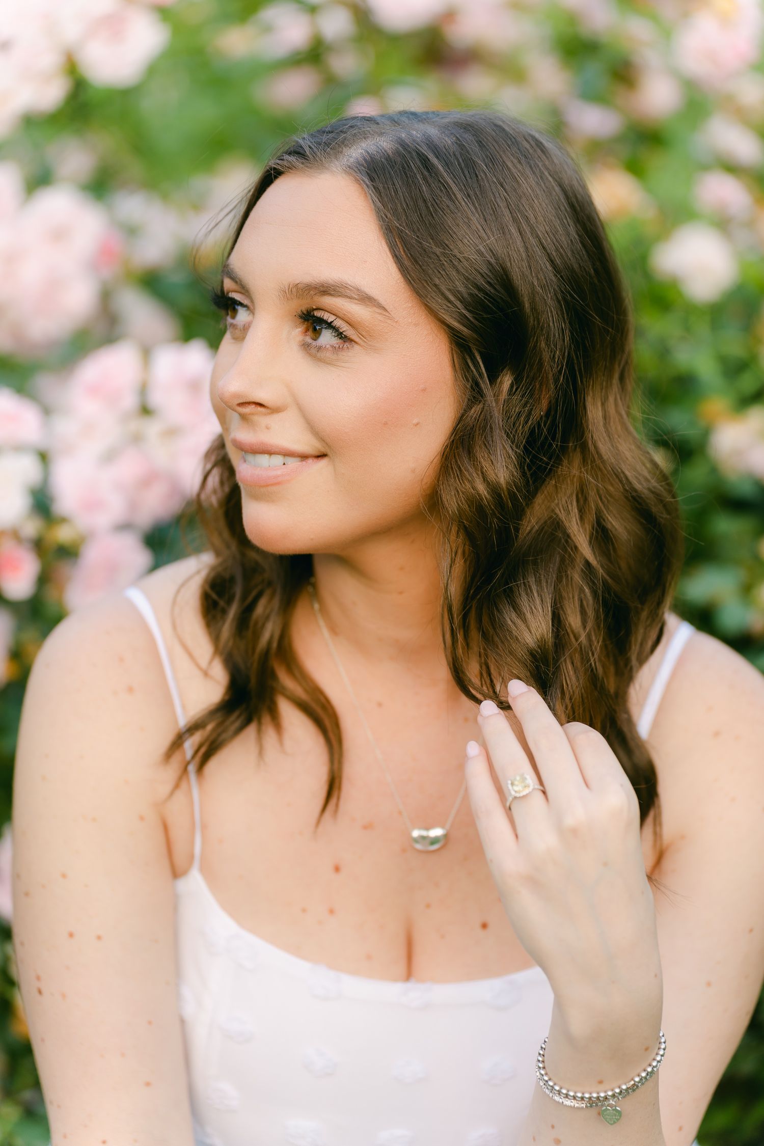 Woman in white dress poses near pink roses showing engagement ring and jewelry.