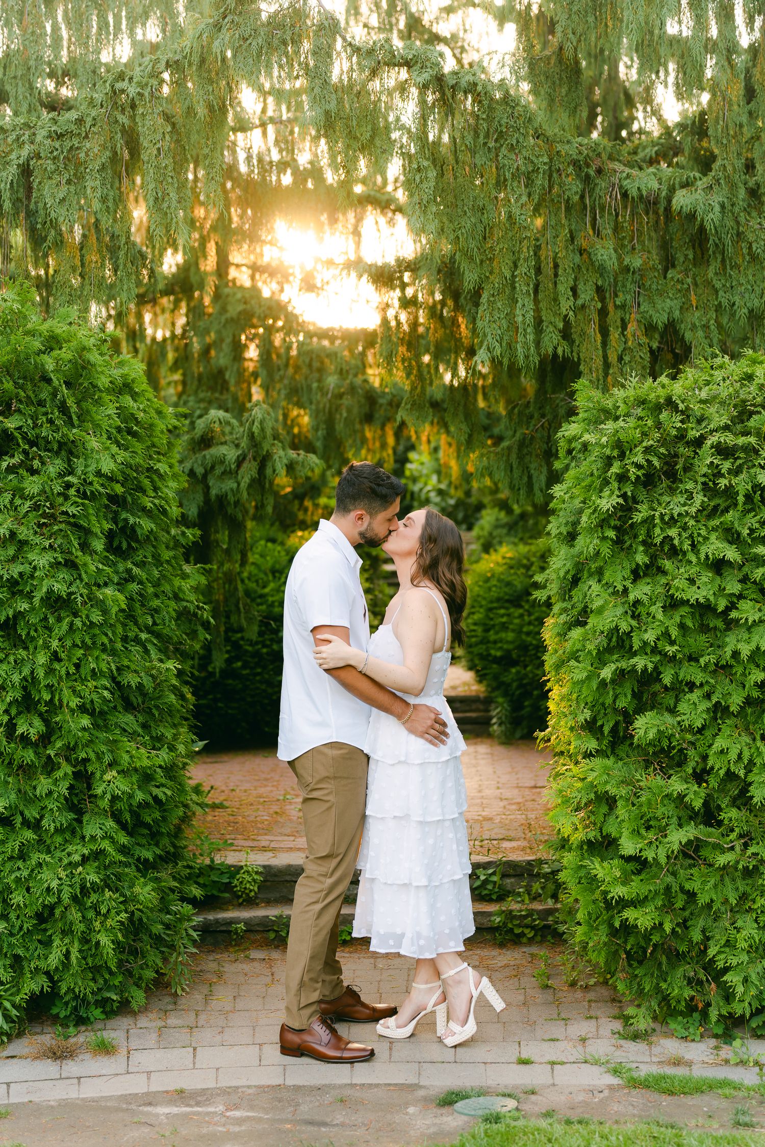 Sunset engagement photo of a couple kissing on stone steps between perfectly trimmed hedges.