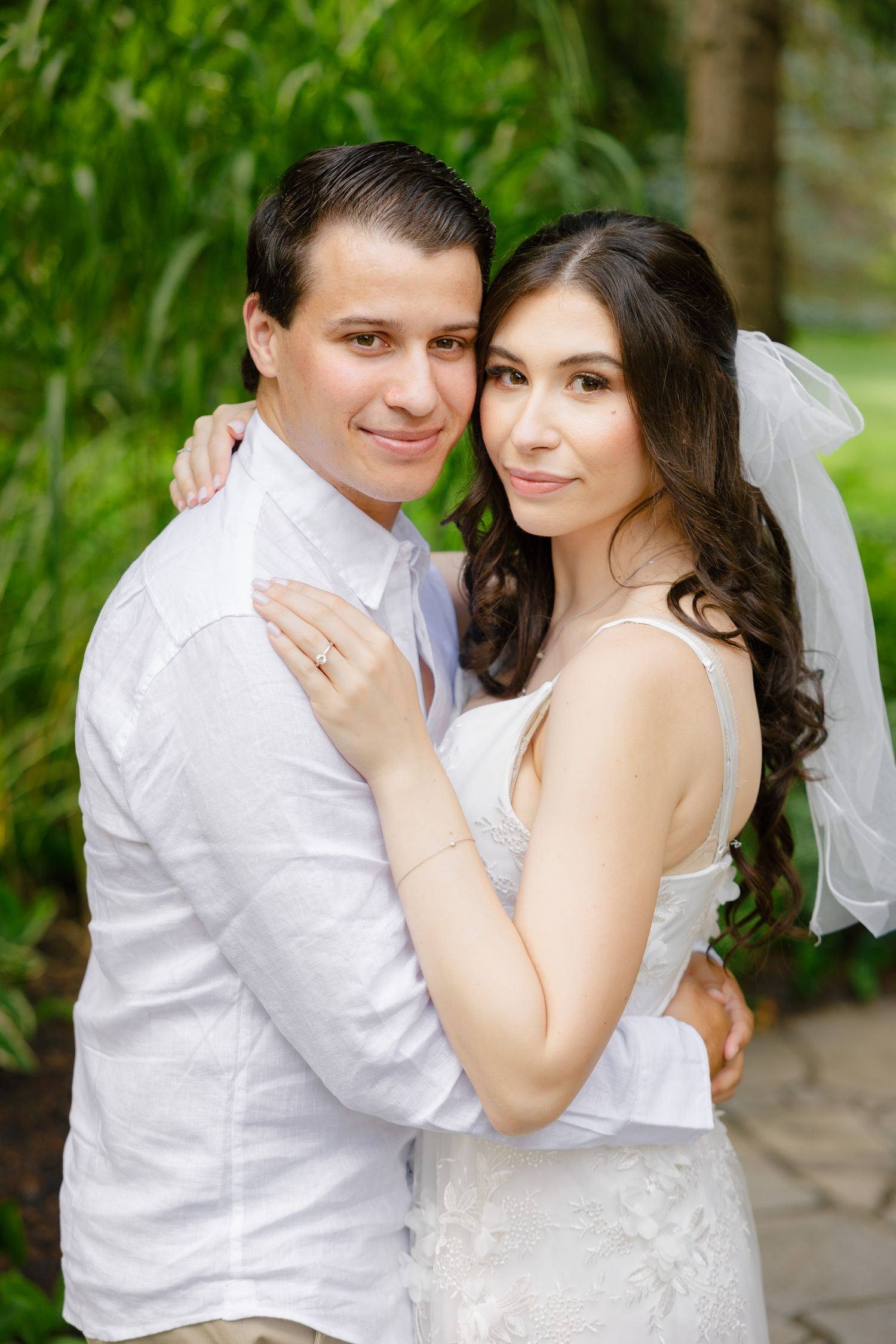 Wedding portrait of couple embracing on garden path with evergreen trees in background.