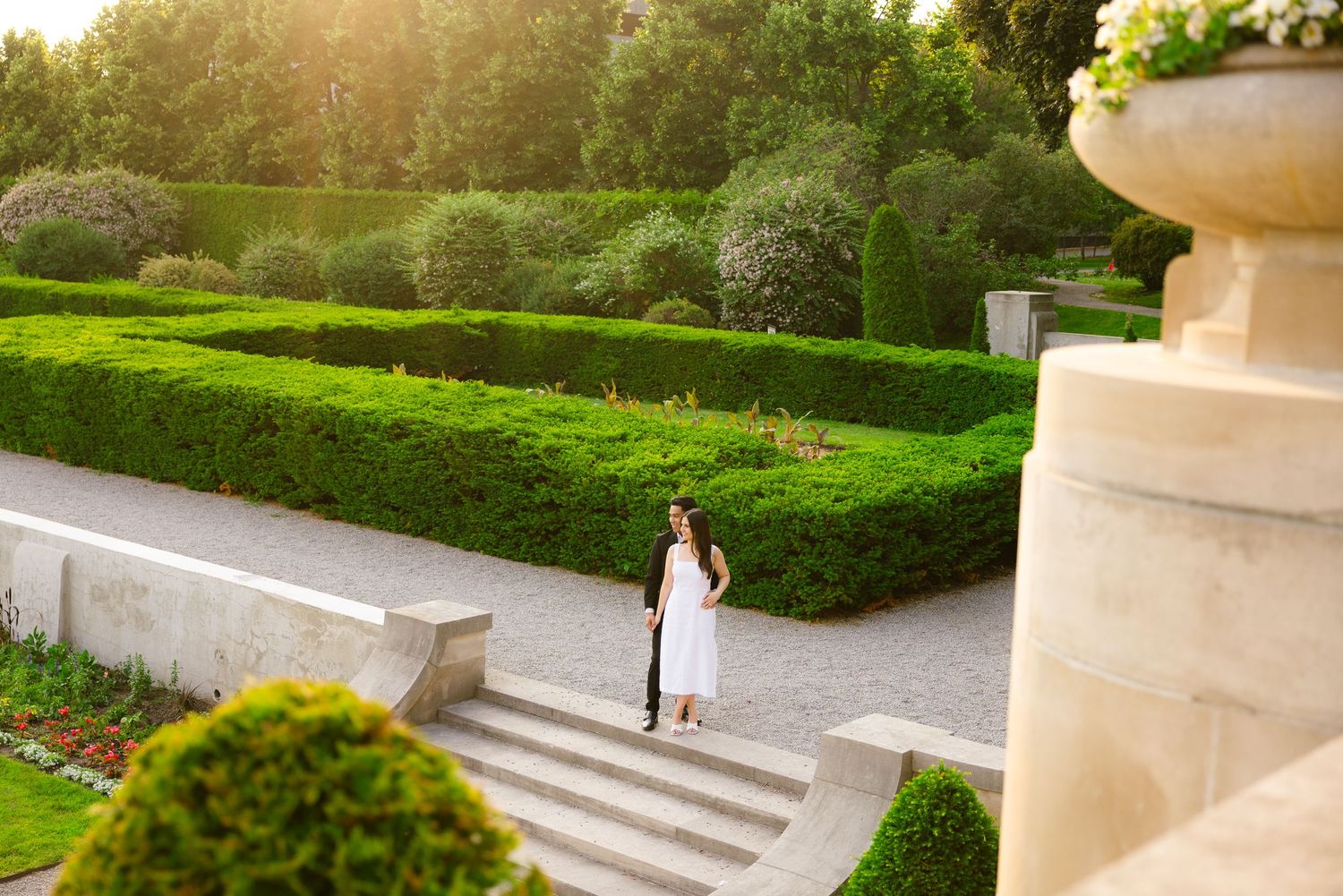A person in white standing on stone garden steps during golden hour with manicured hedges in the background.