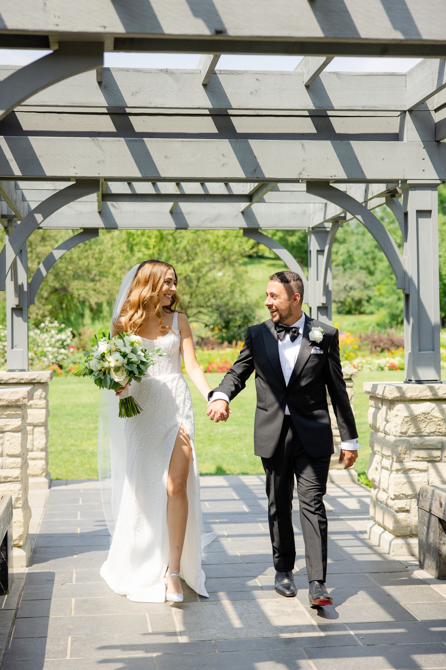 Joyful wedding couple walking together under an elegant stone and wood pergola.