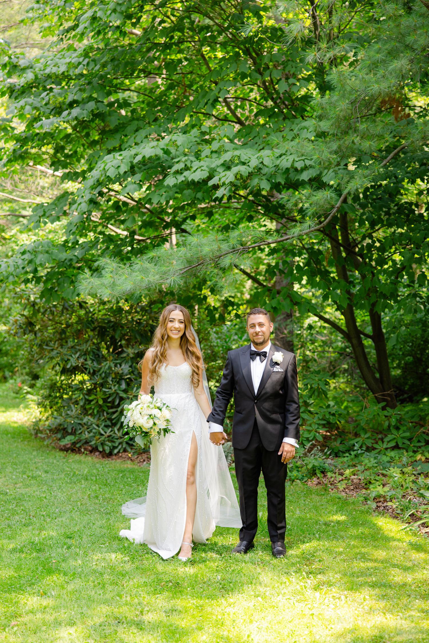Wedding couple poses together on grass with leafy green trees creating a natural backdrop.
