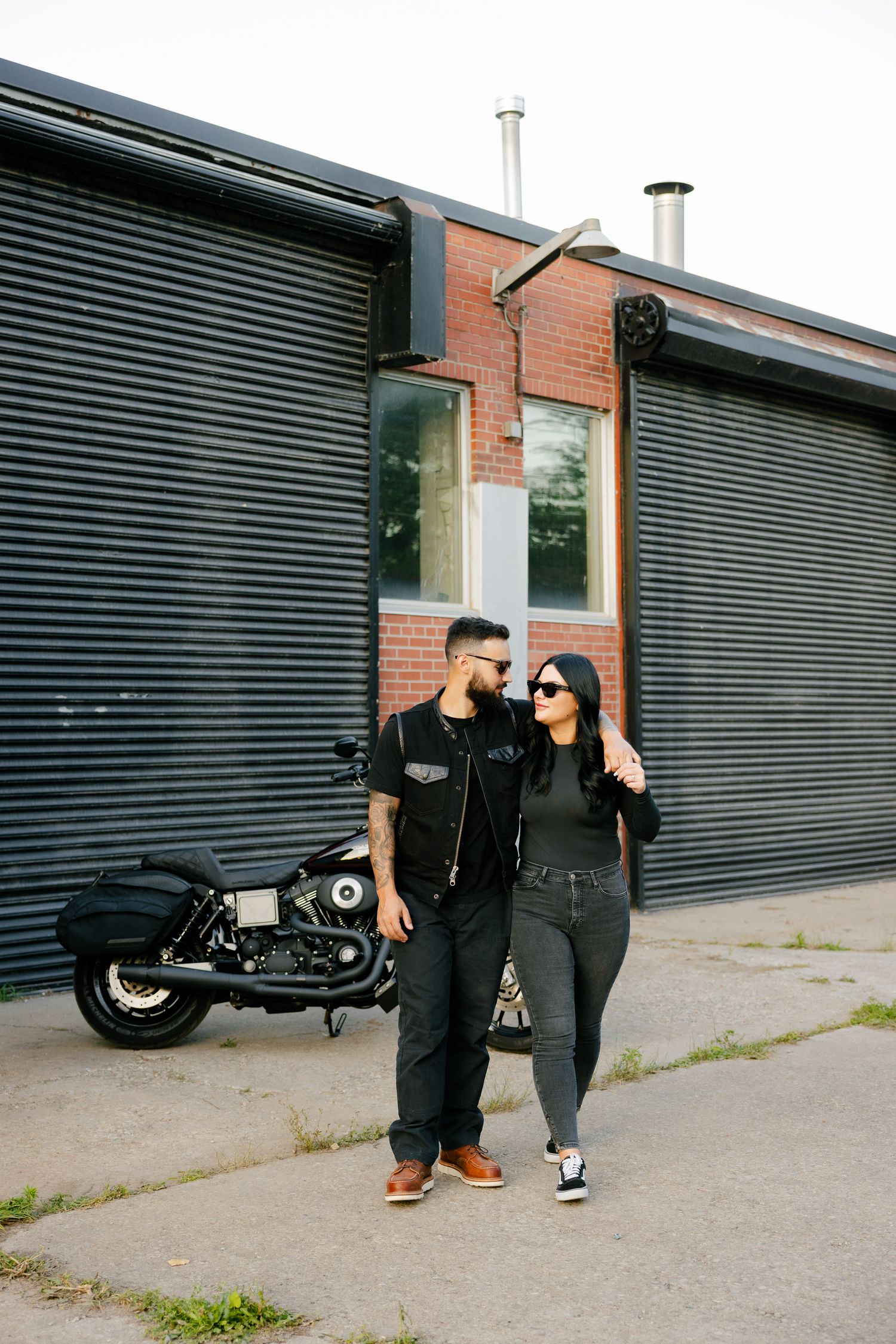 Couple in matching dark outfits stands near motorcycle by brick building.