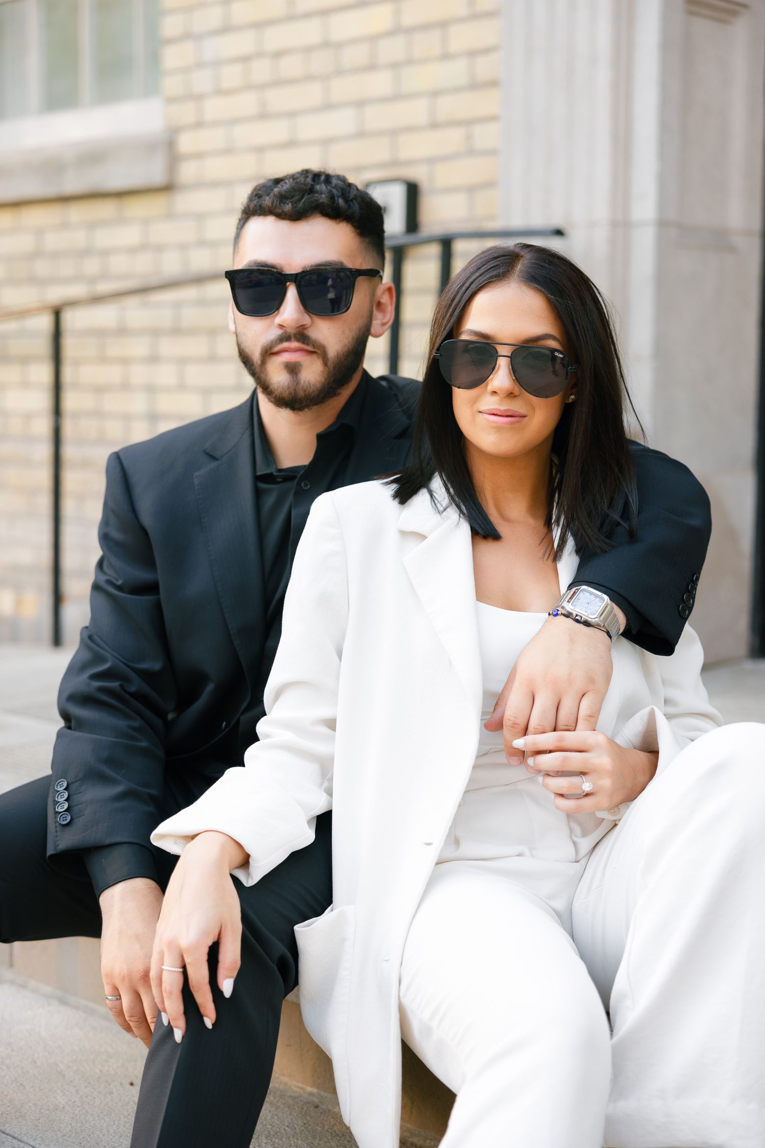 Chic couple in sunglasses and coordinated formal wear pose against brick building.