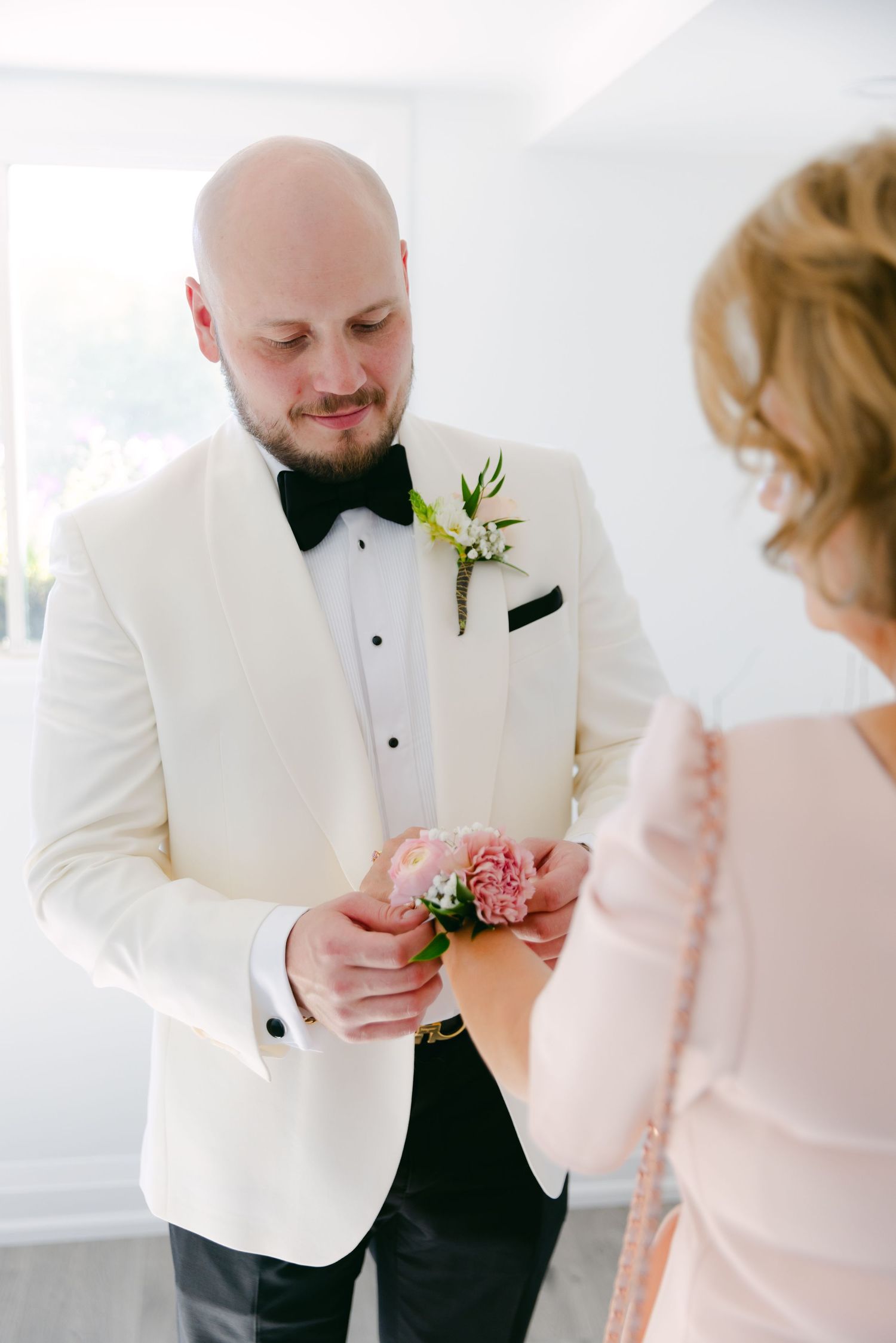 A groom in white tuxedo receives a pink flower corsage during wedding preparations.
