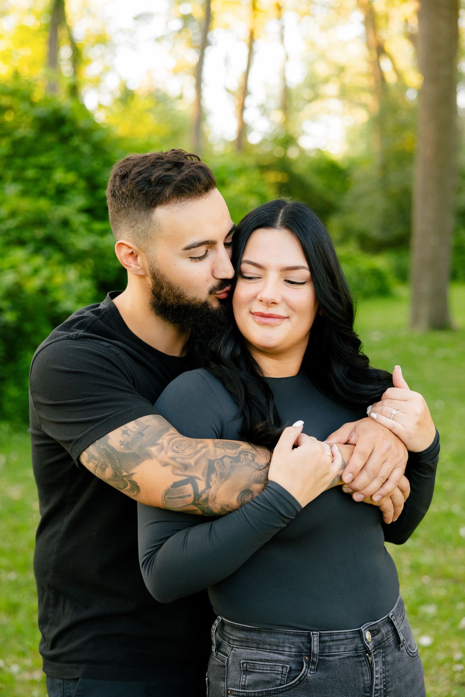 Romantic outdoor portrait of couple in black clothing embracing against sunlit forest backdrop.