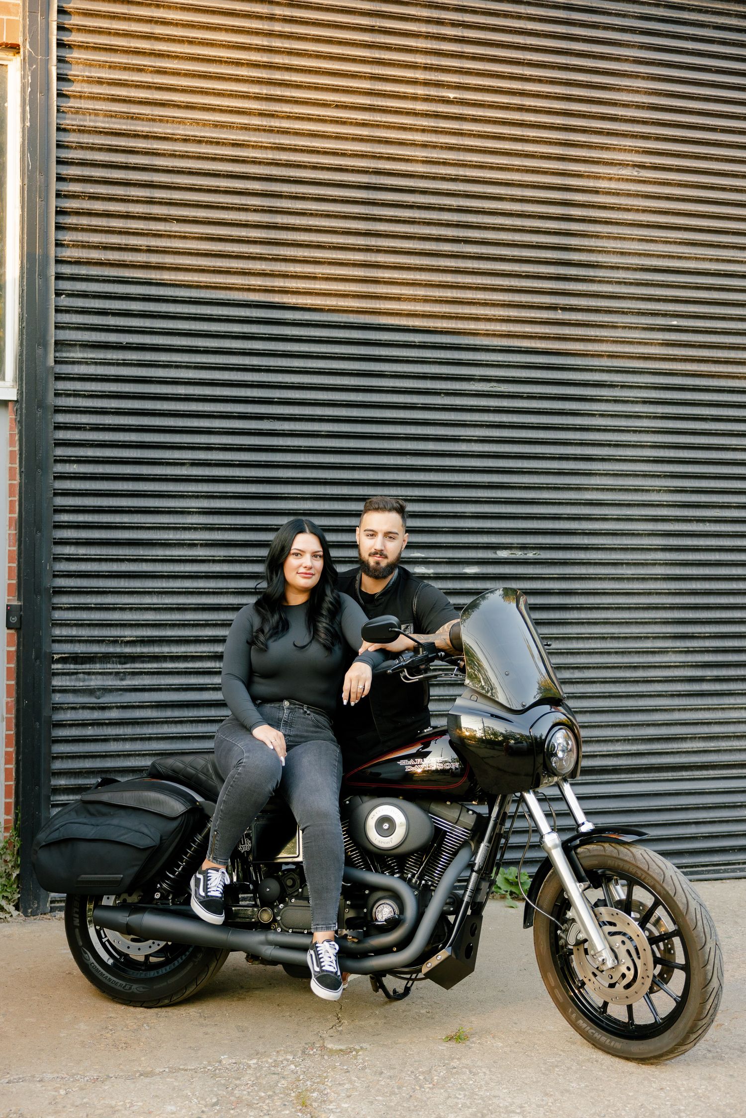 Two people sitting together on a custom motorcycle with industrial background.