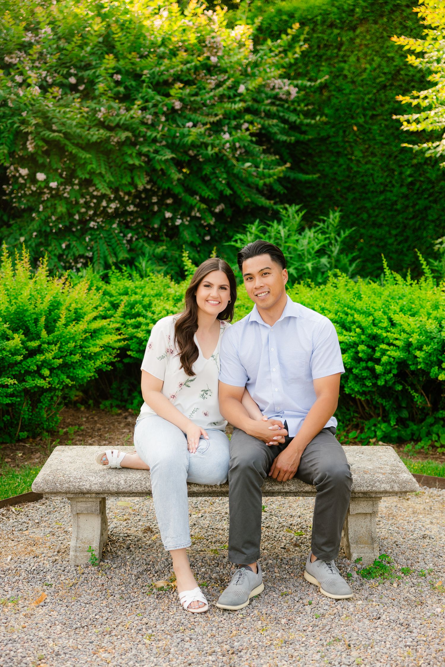 A happy couple poses together on a stone bench surrounded by manicured garden shrubs.