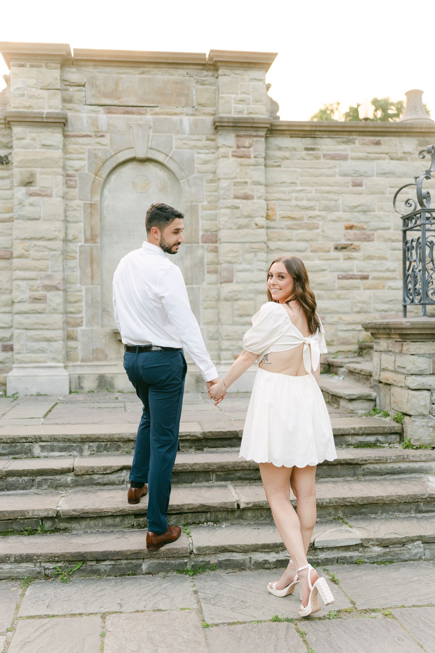 A couple in coordinated white outfits holding hands while walking up stone steps with decorative iron railings.