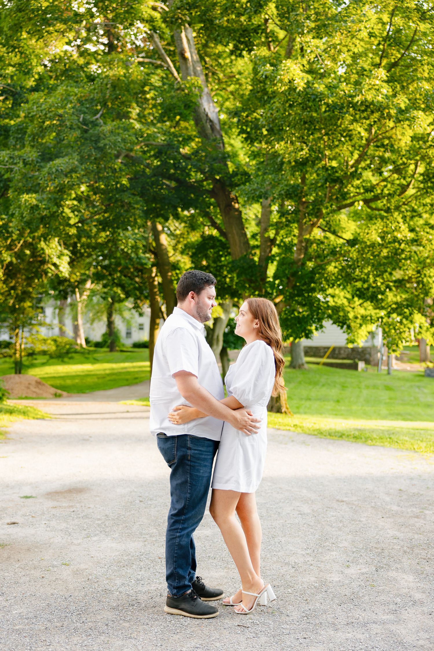 A couple in matching white outfits embrace on a walking path surrounded by green trees.