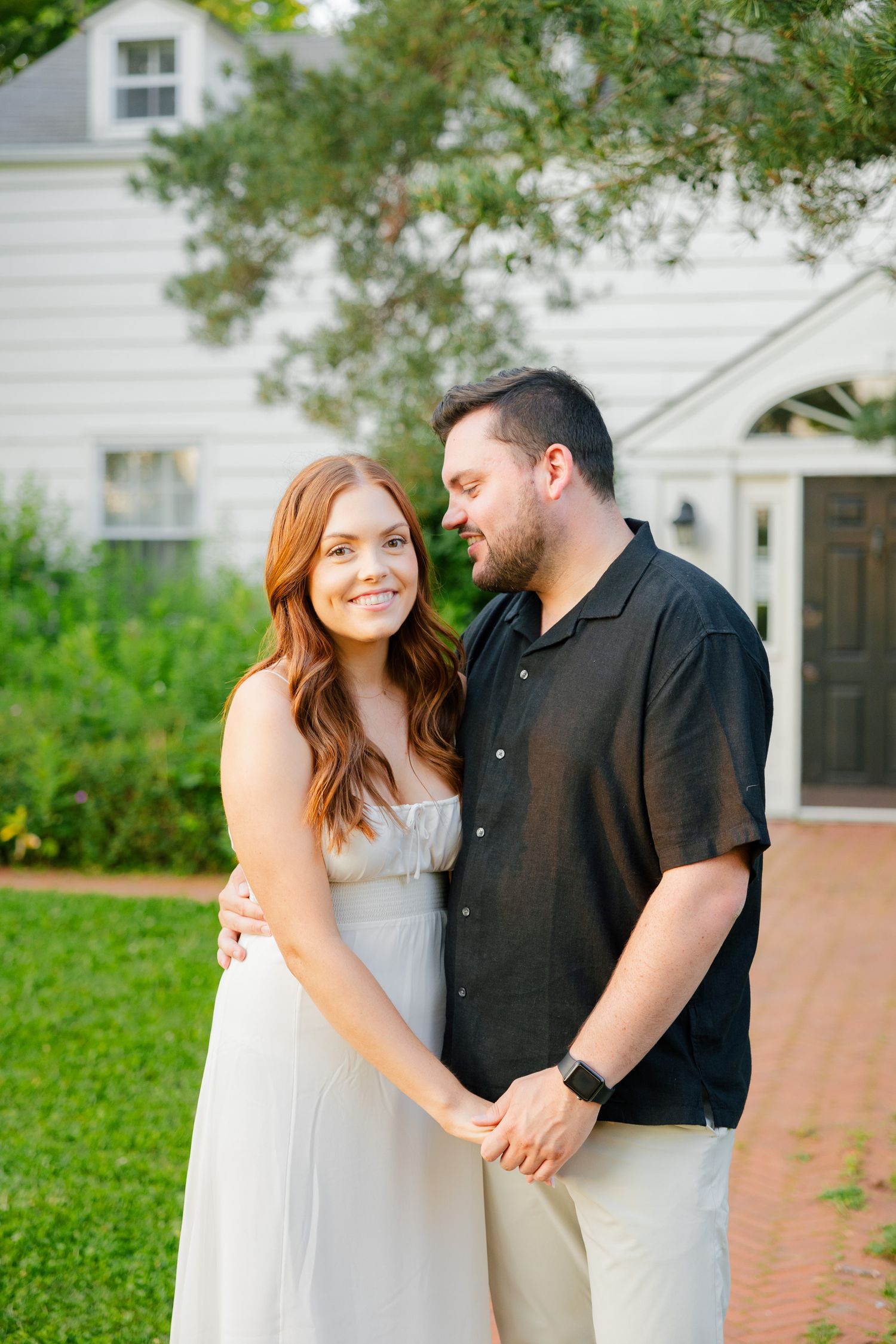 Couple shares an intimate moment in a garden setting with a white house backdrop.