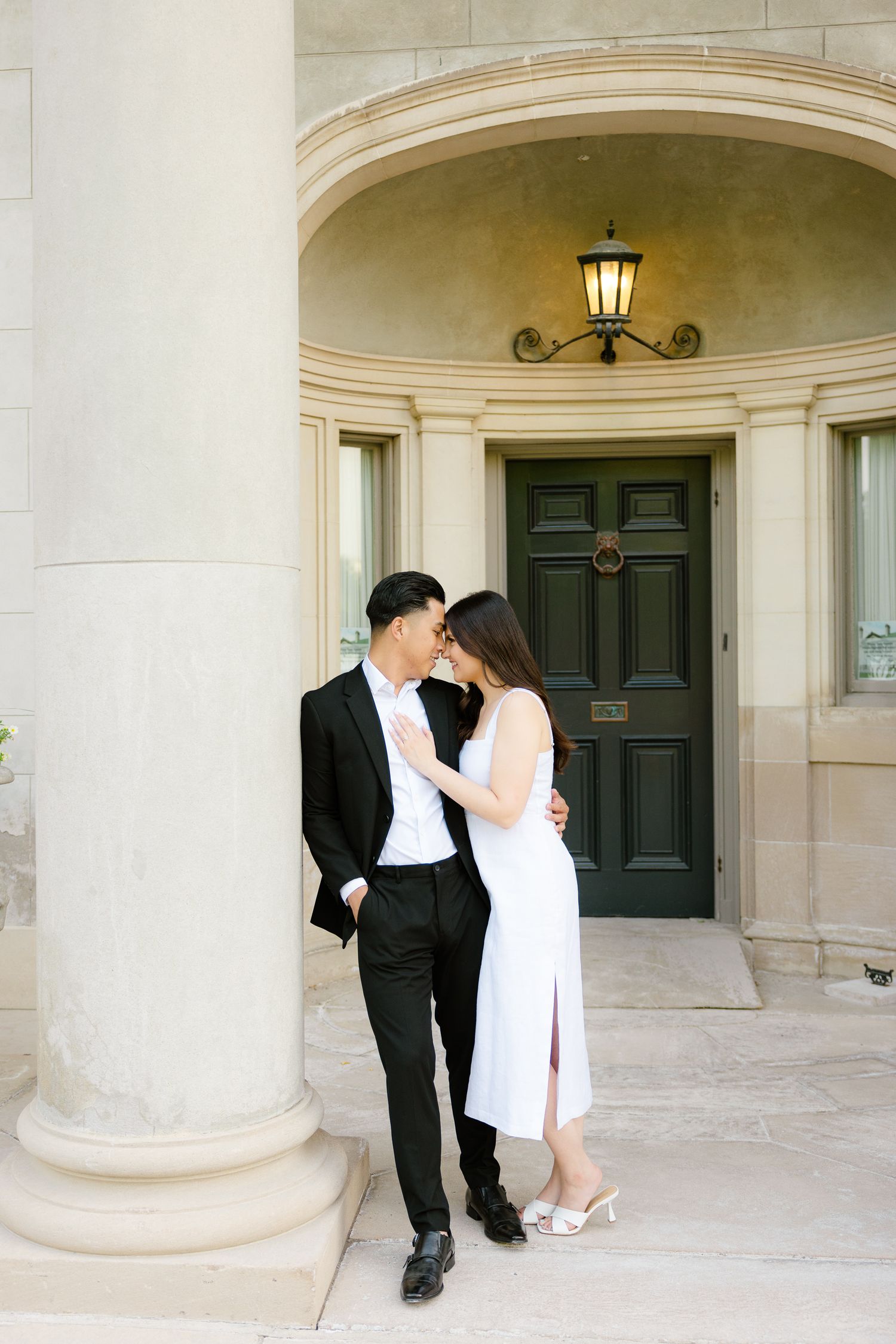 Romantic couple in formal attire pose intimately by grand entrance with vintage lamp.