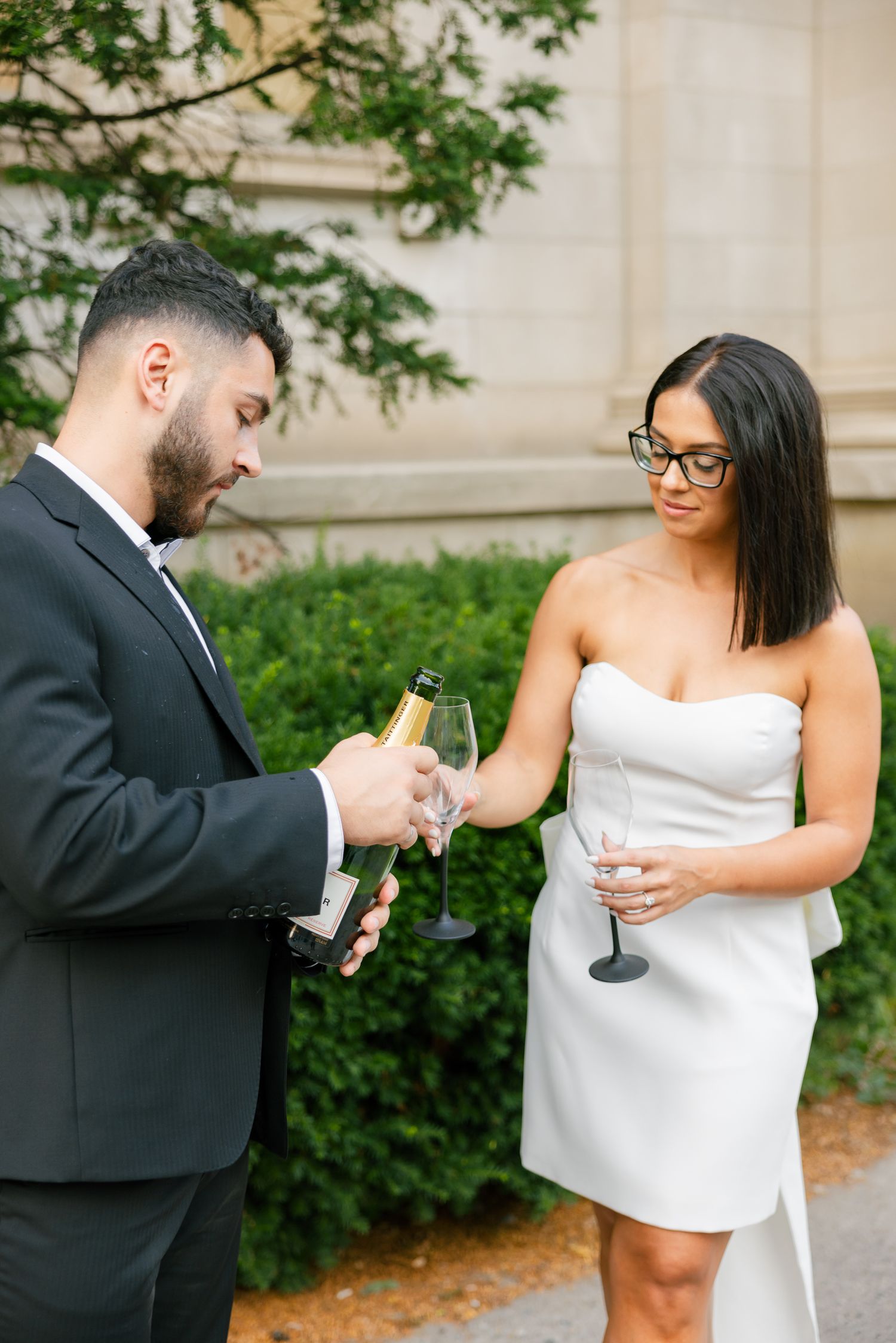 A couple pours champagne into glasses while standing beside manicured bushes and stone architecture.
