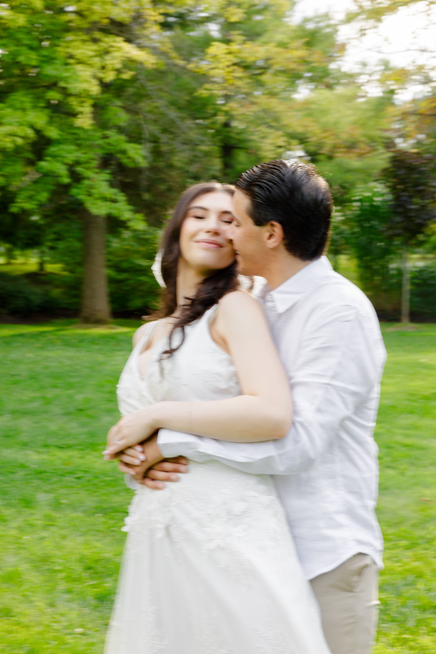 Motion-blurred candid photo of couple in white embracing outdoors with natural greenery.
