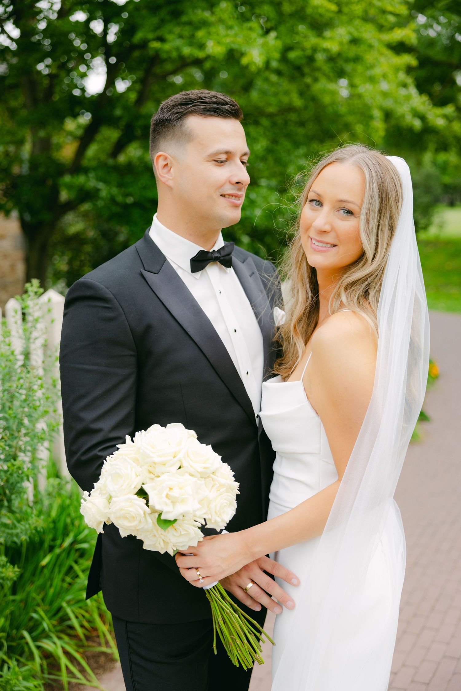 Elegant couple in wedding attire poses outdoors with a white bouquet and garden backdrop.