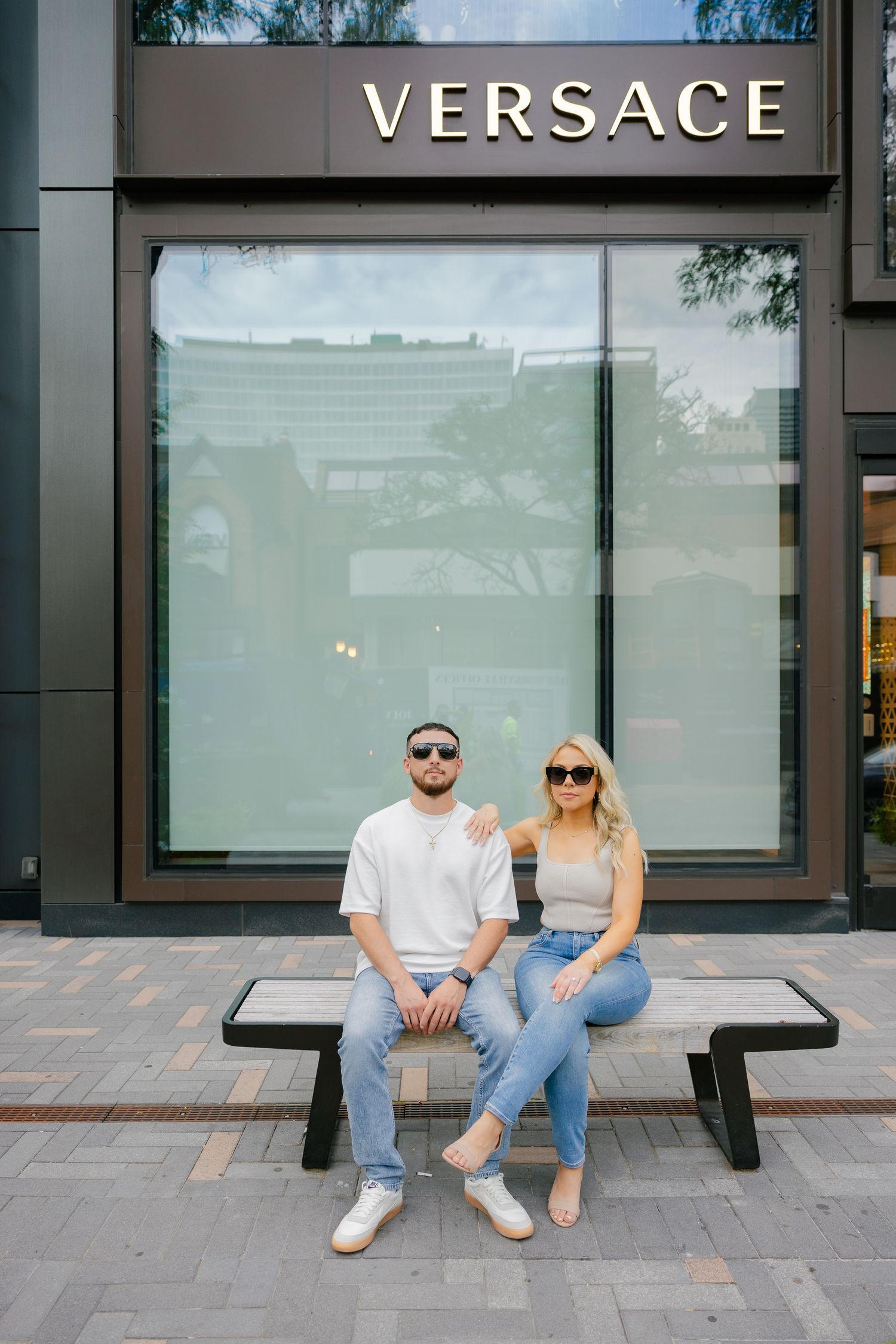 Couple sitting on bench outside Versace store window in matching casual attire.