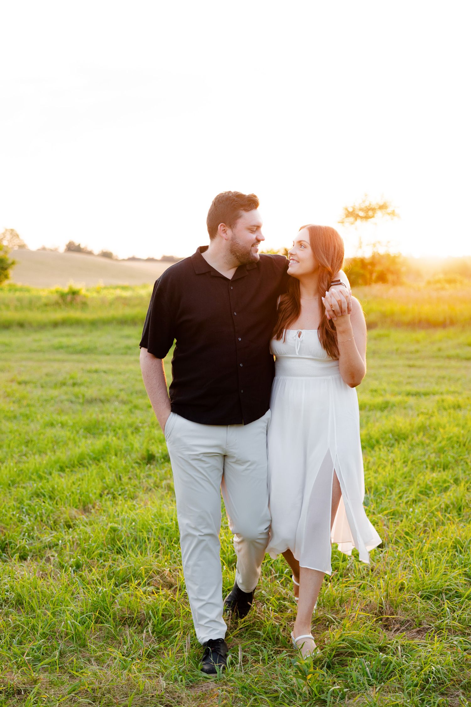 Couple walks hand in hand through sunlit field during summer engagement session.