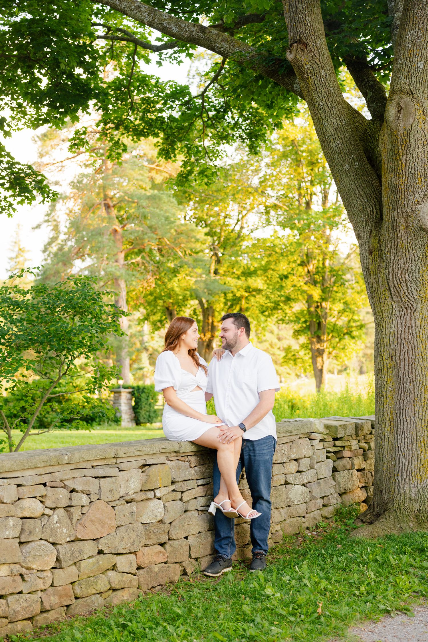 A couple shares a romantic moment on a rustic stone wall beneath a large tree in a sunlit garden setting.
