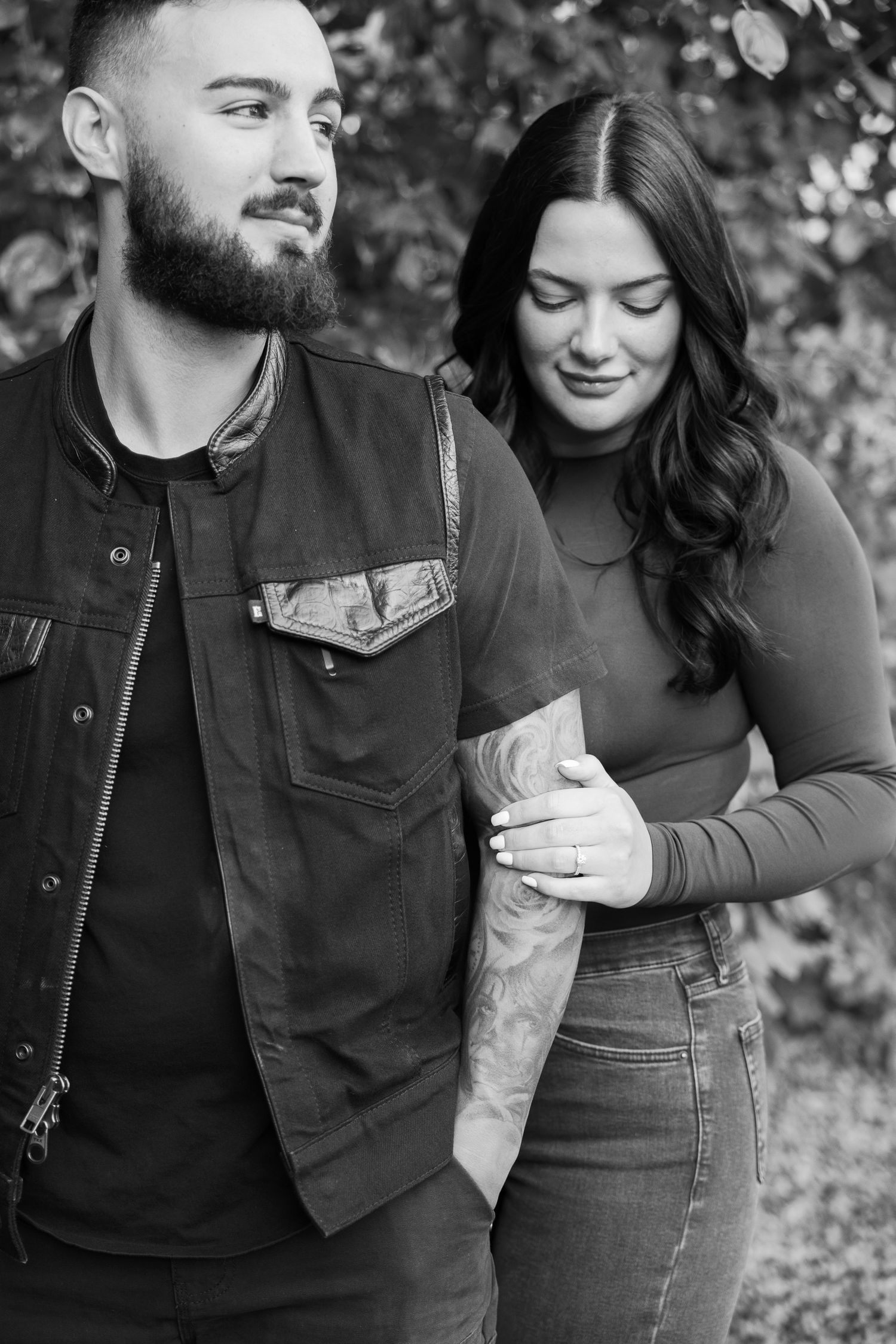 A black and white portrait of a couple in casual wear standing close together outdoors.