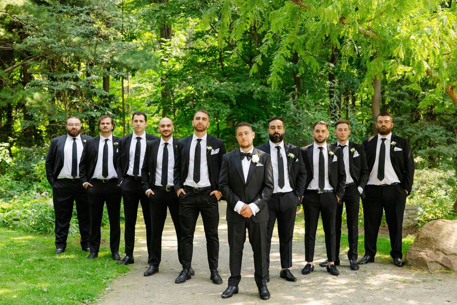 A wedding party of groomsmen in black suits and ties pose together on a garden path surrounded by greenery.