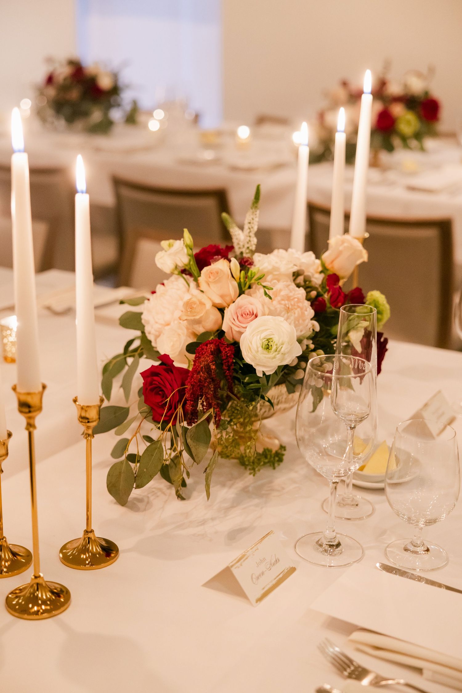 Wedding table setting with blush and burgundy floral centerpiece and brass candlesticks on white linens.