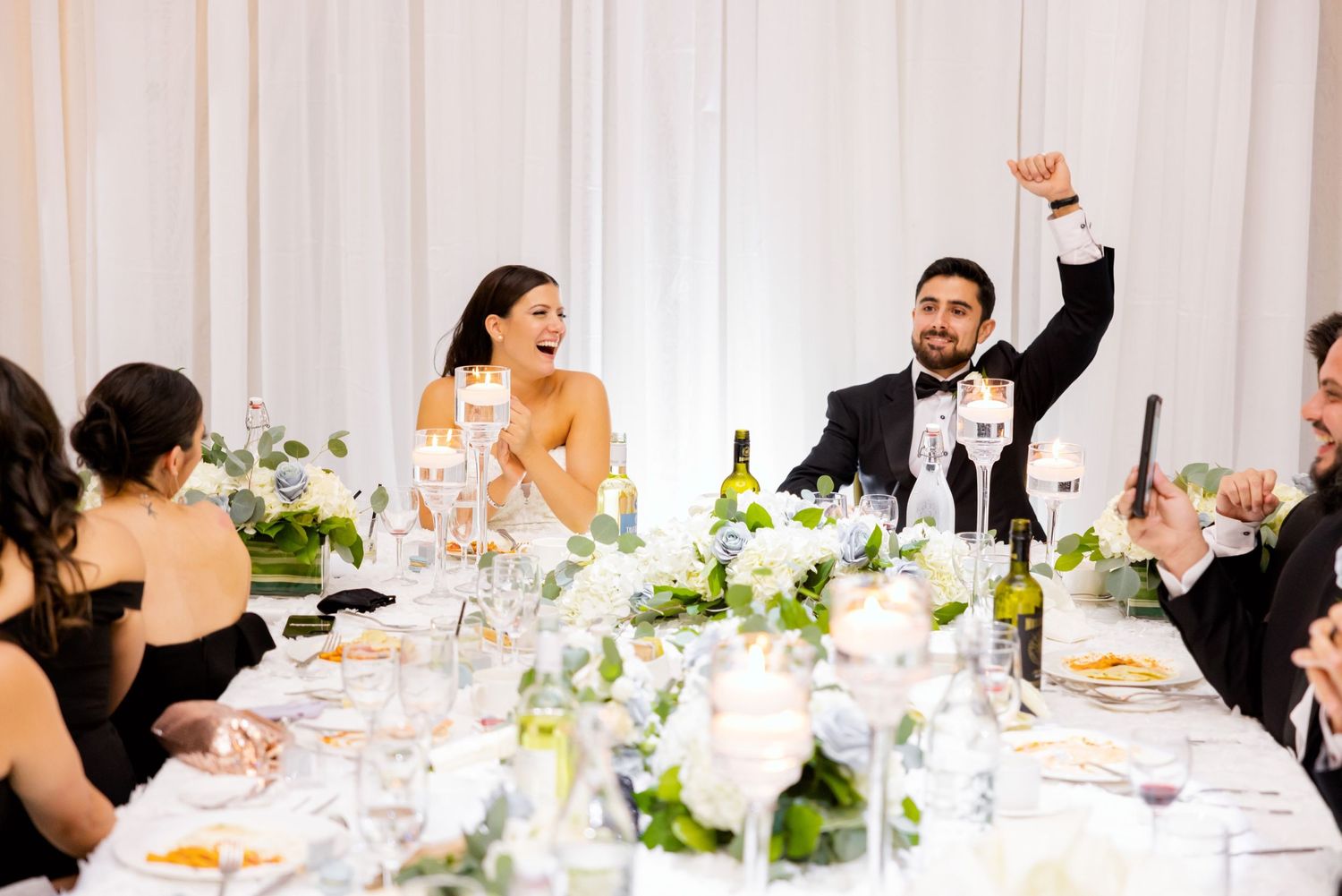 Guests at wedding reception table raising glasses in celebration with white floral decorations.