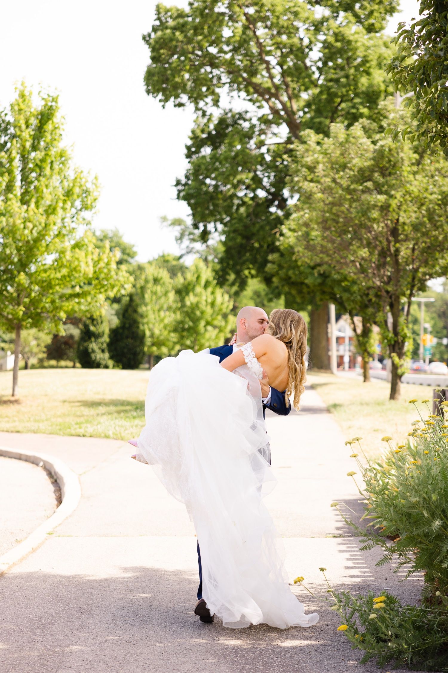 Wedding couple embraces on a tree-lined path with flowing white dress.