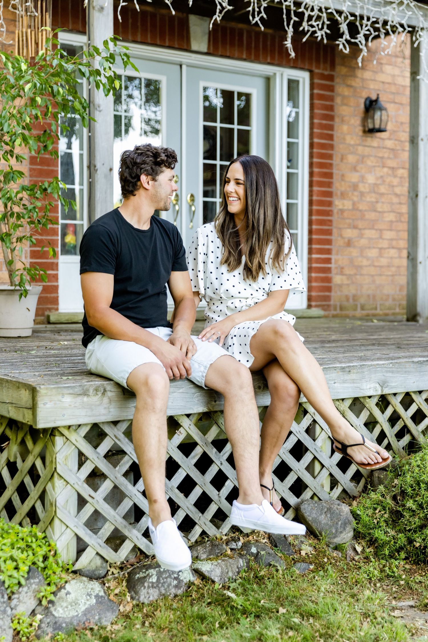A couple in summer attire sits intimately on a wooden deck lattice outside a brick house with white French doors.