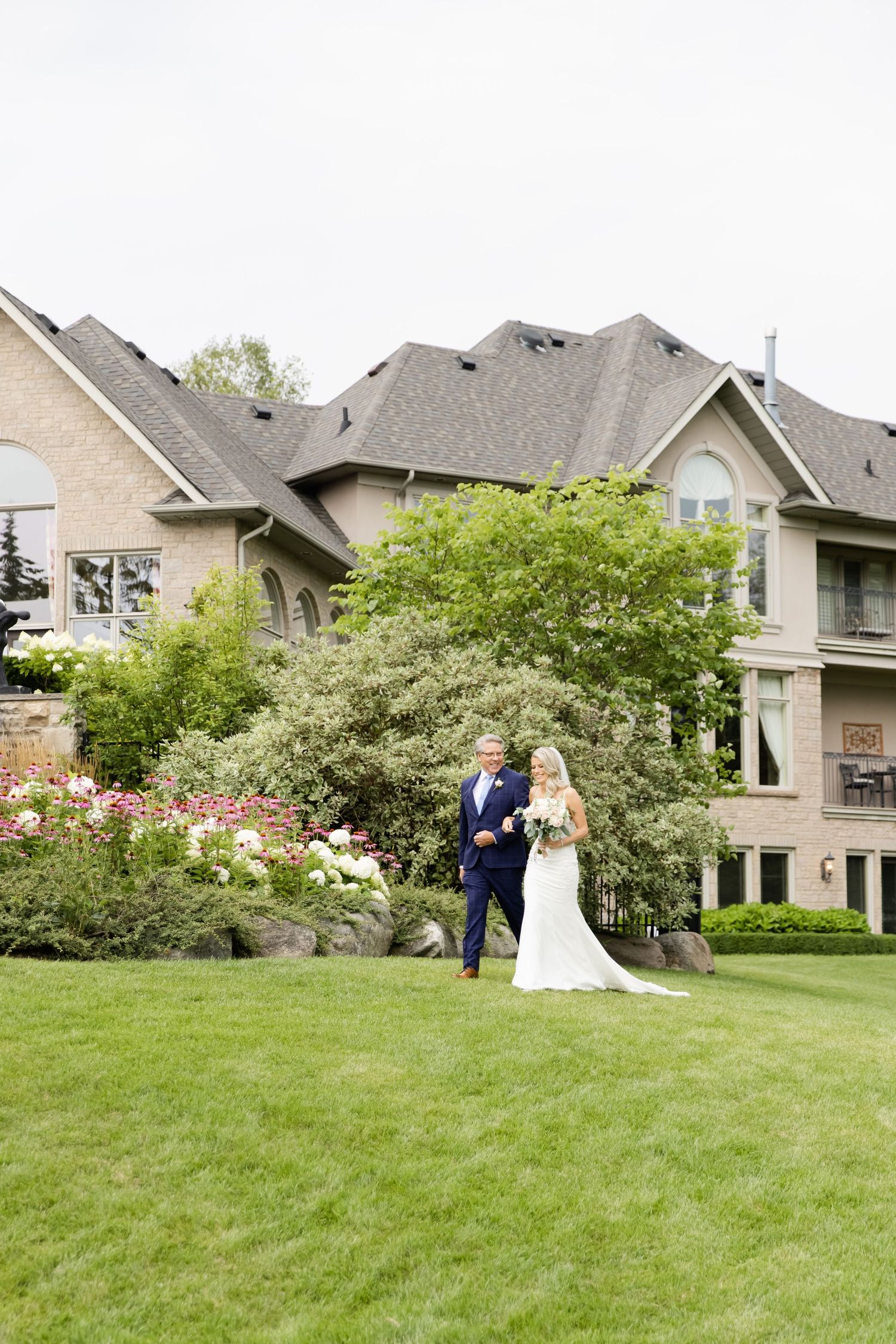 Wedding couple poses on lawn in front of luxurious multi-level home with landscaped garden.