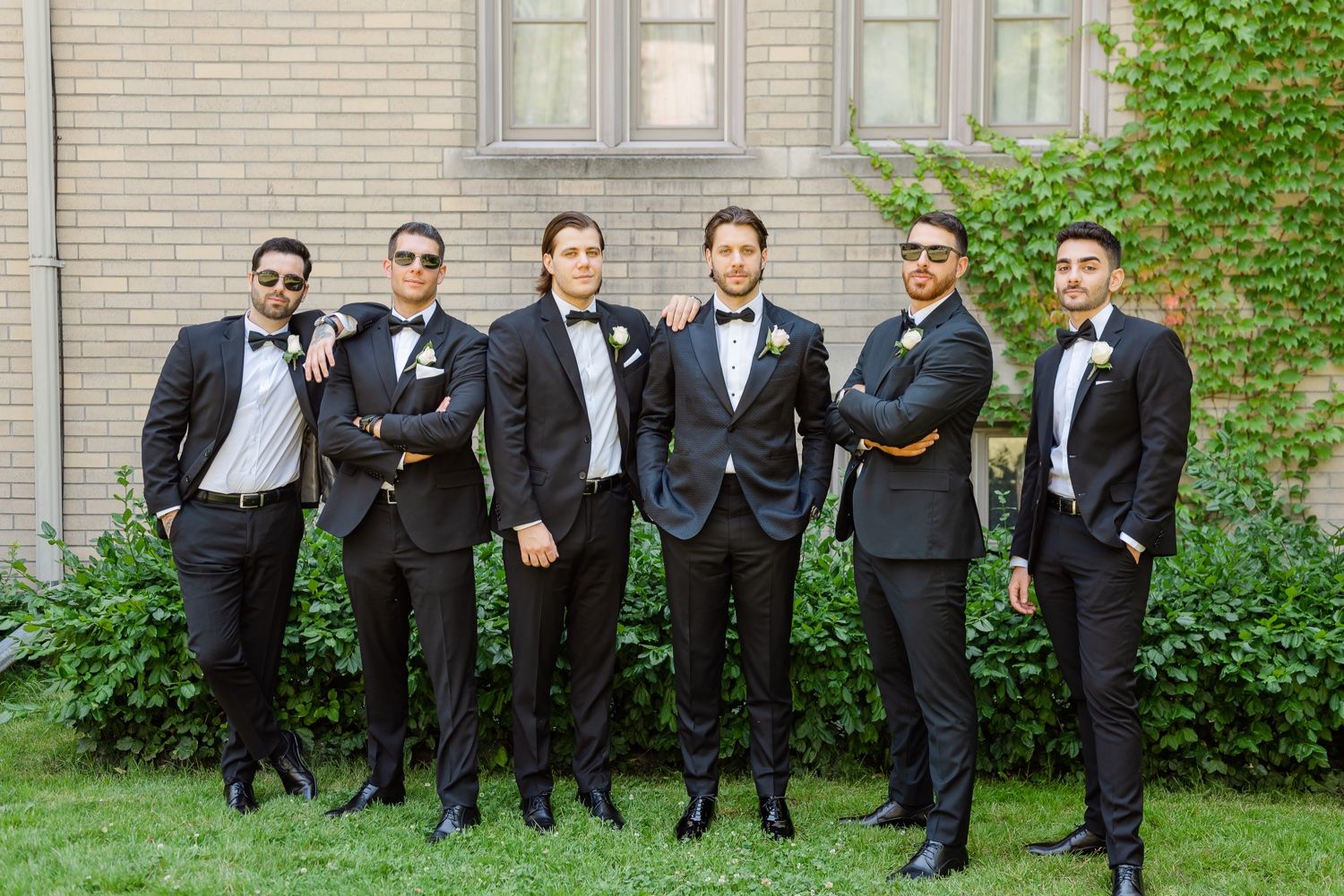 Wedding party groomsmen in black tuxedos stand together against brick wall with greenery.