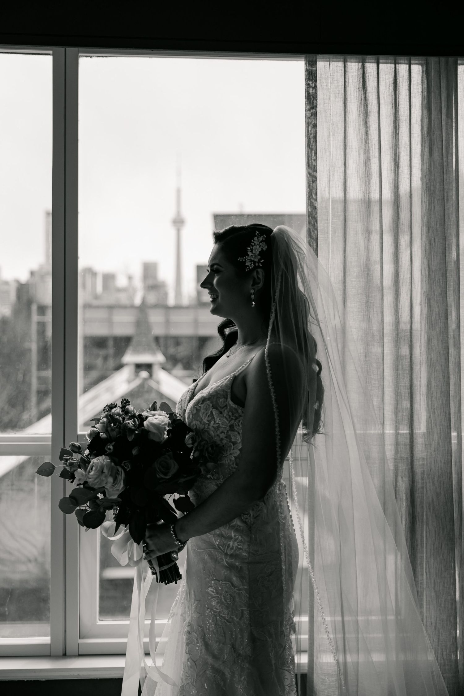 Black and white silhouette of bride holding bouquet against window with city skyline backdrop.