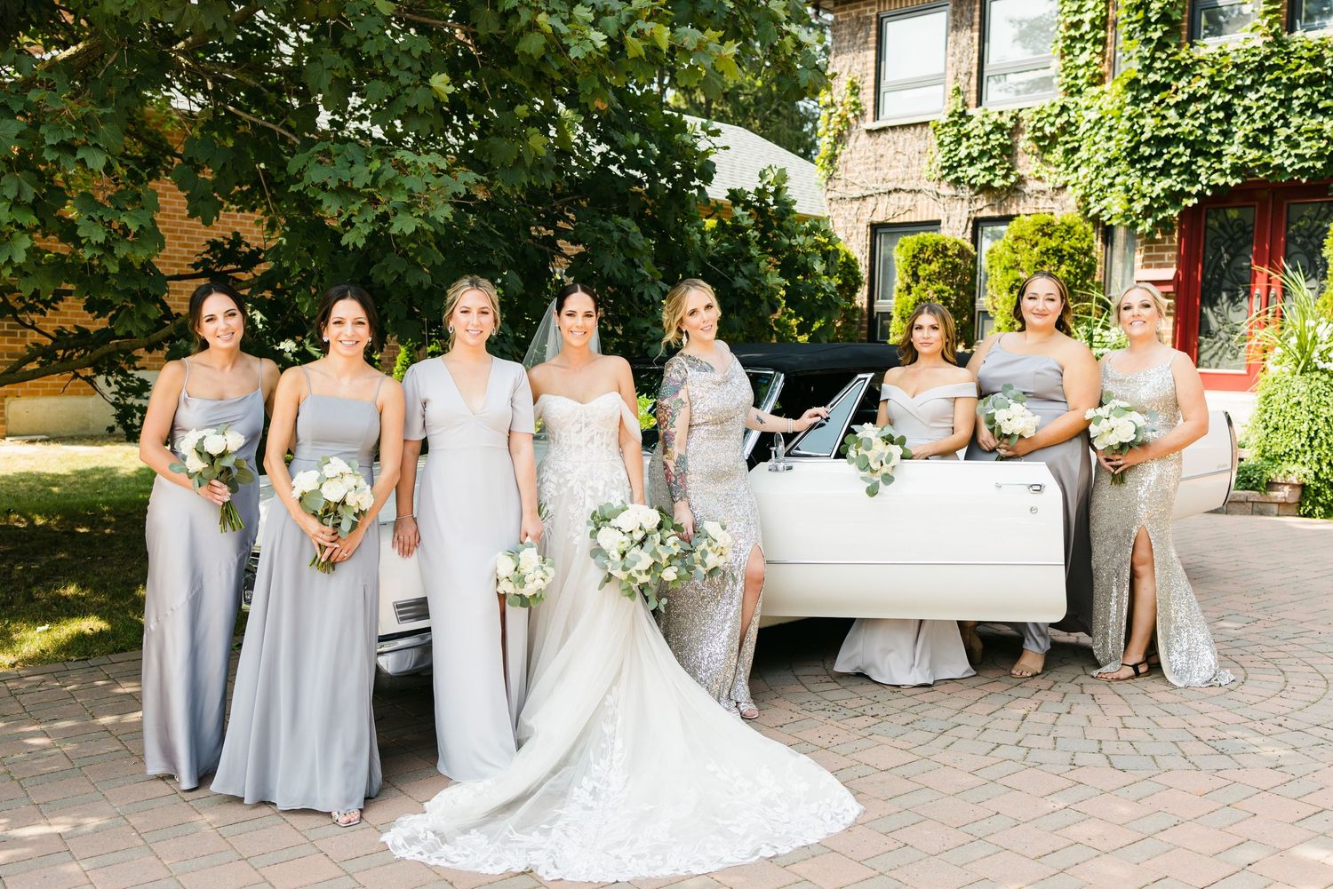 Wedding party in silver bridesmaid dresses posing with a classic white car on a brick courtyard.