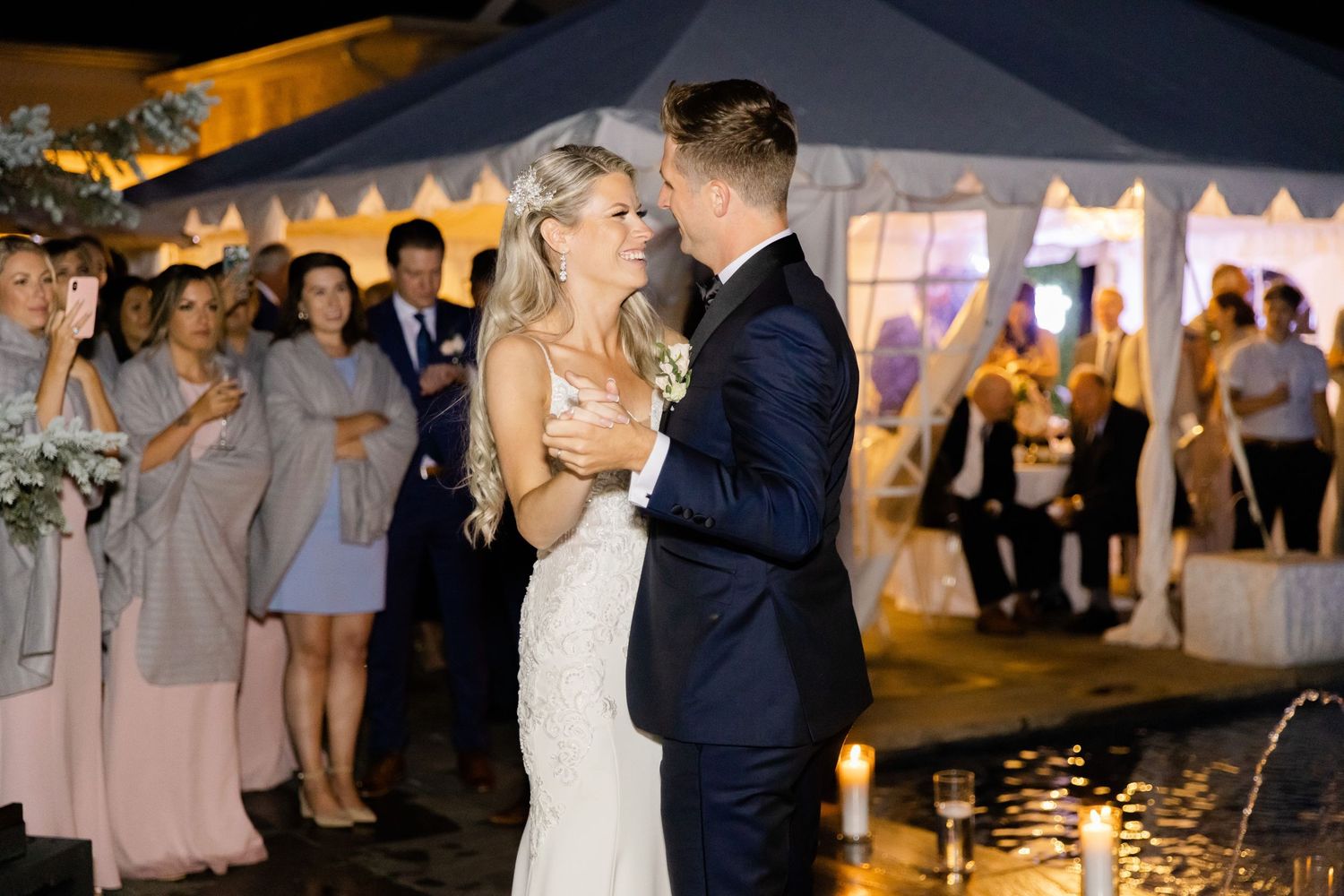 Evening wedding celebration with couple dancing surrounded by guests under tent lighting.