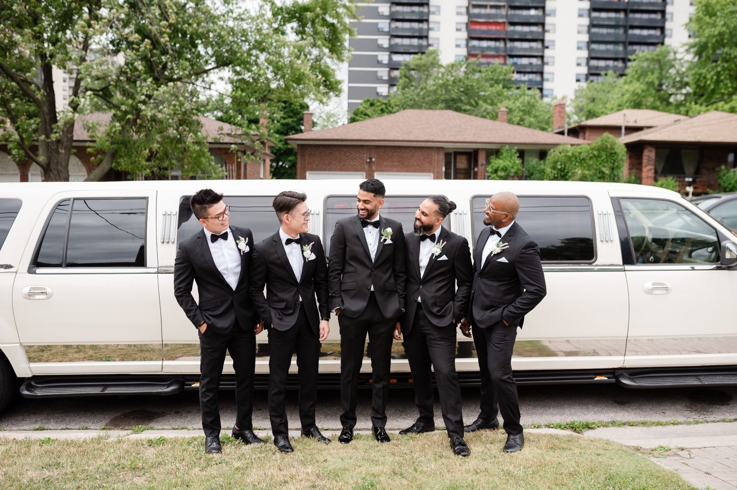 A group of groomsmen in black tuxedos pose together in front of a white stretch limousine.