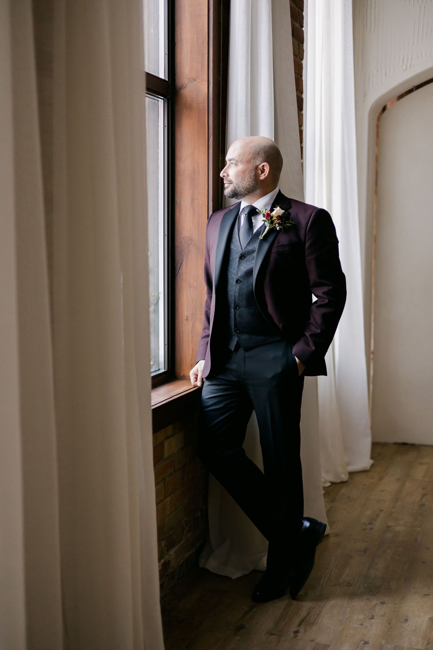 A groom in formal attire leans against a window frame in contemplative pose.