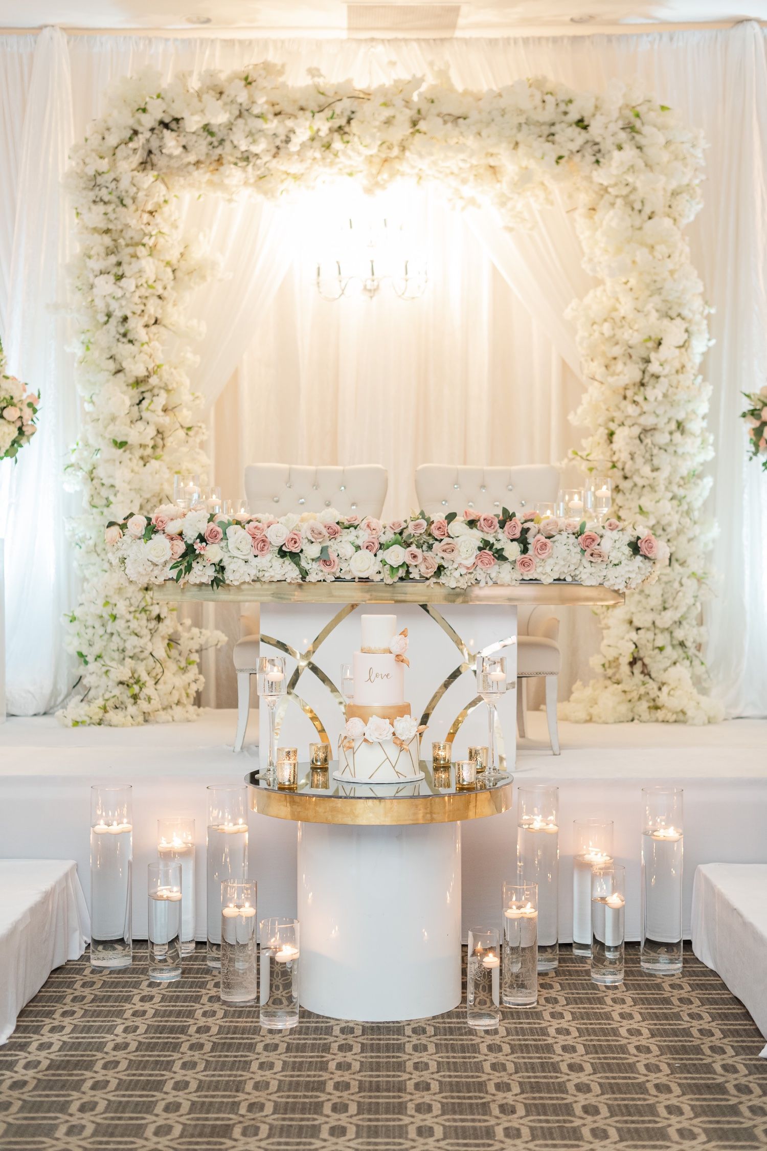 Romantic wedding stage with white floral arch, candles, and elegant cake display in soft lighting.