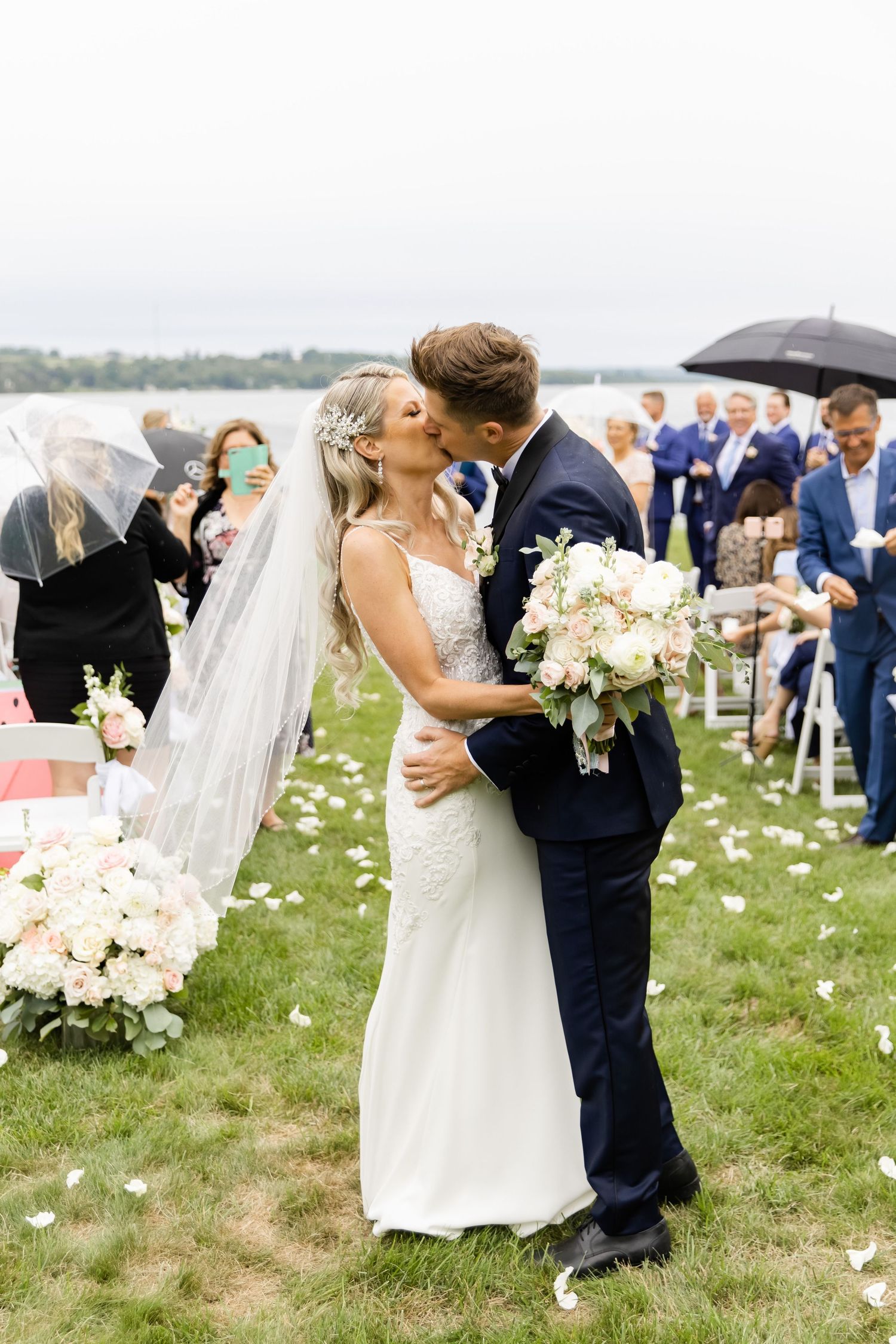 Romantic kiss during outdoor wedding ceremony with flower petals and guests.