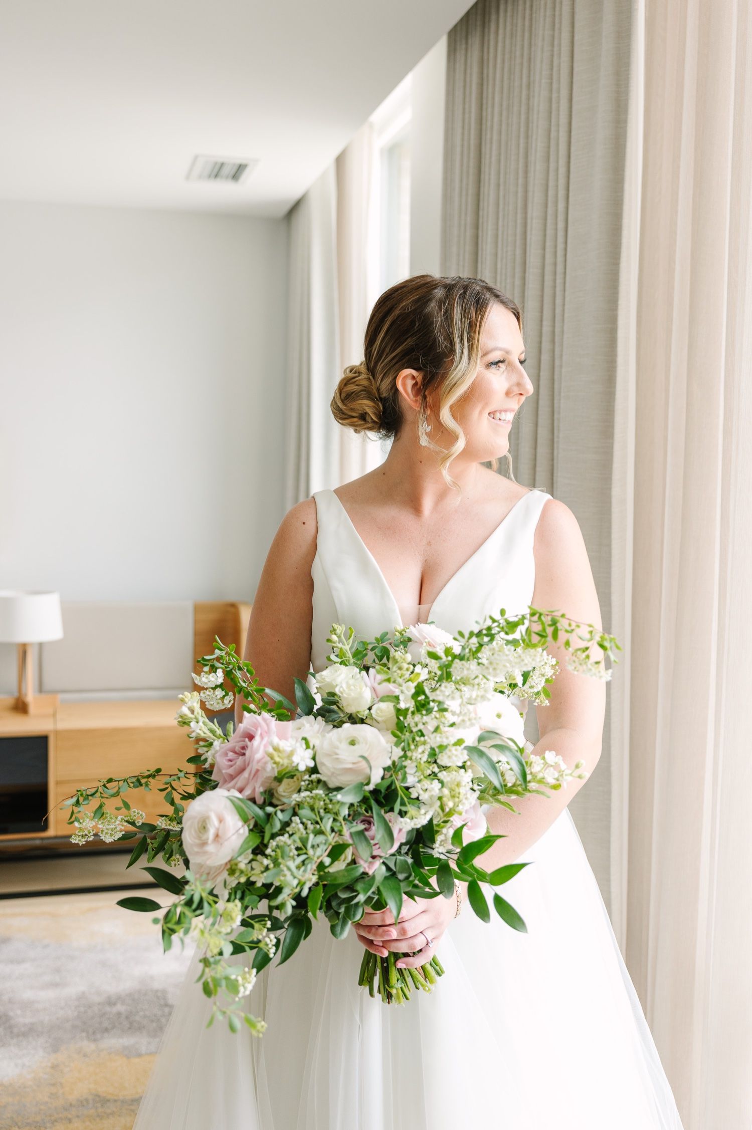 Bride in classic white gown poses by window with fresh spring bouquet.