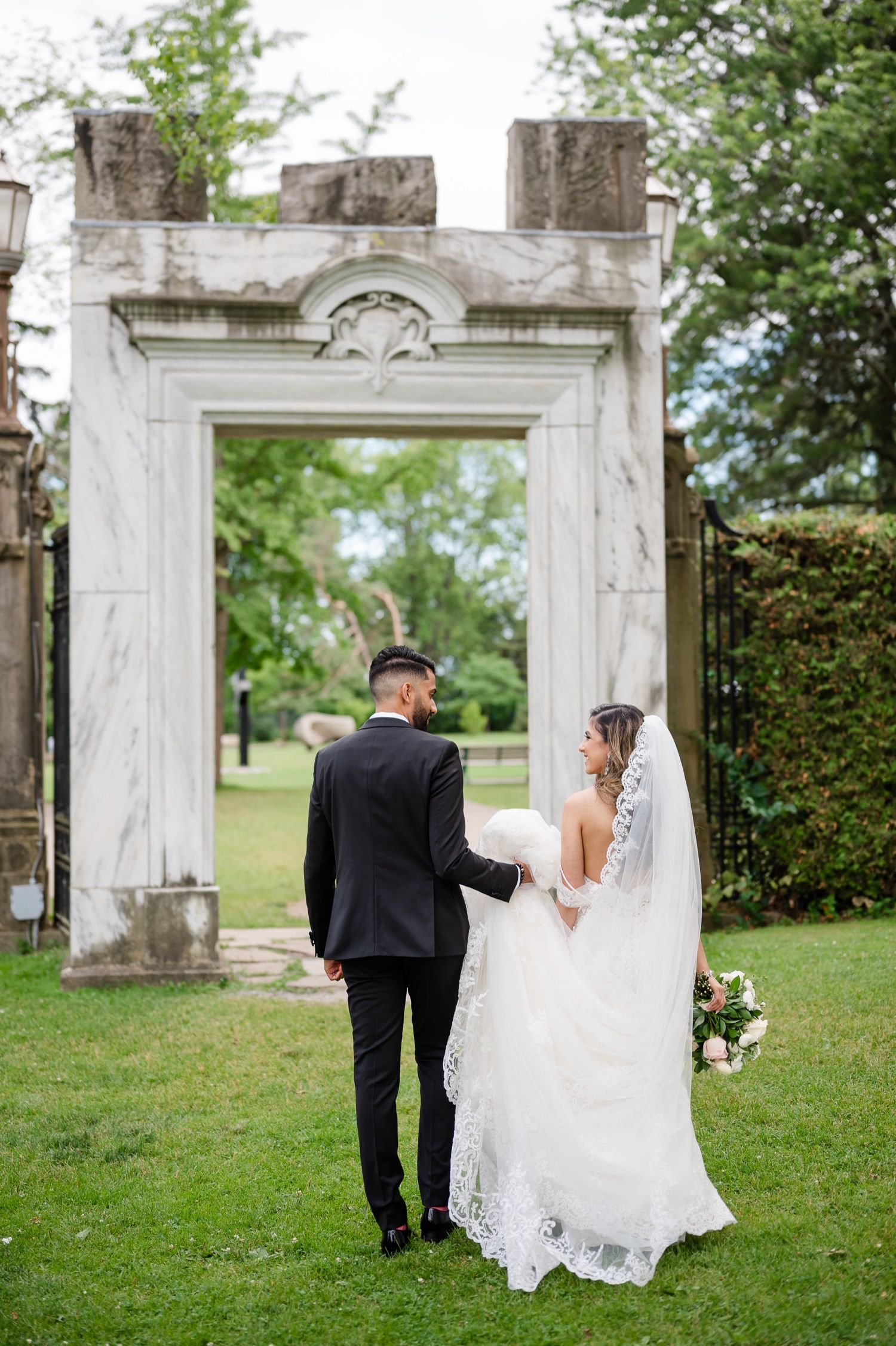 Back view of newlyweds standing before an ornate stone archway in a garden setting.