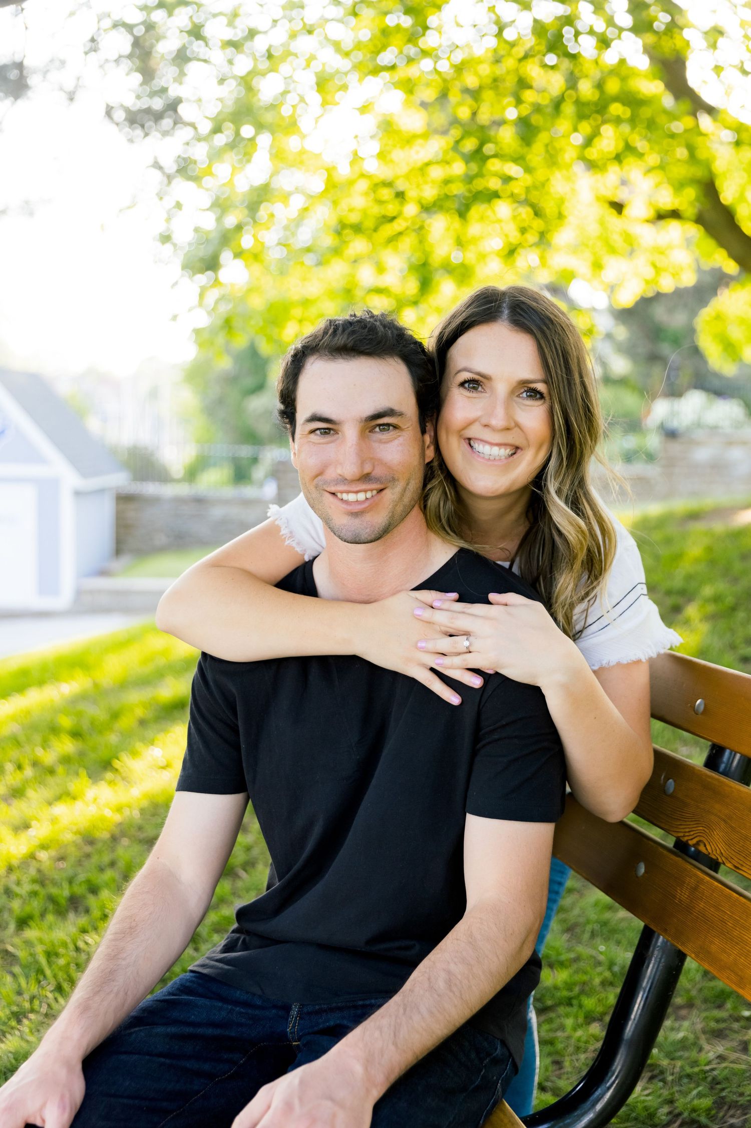 A couple poses together on a park bench with beautiful golden sunlight filtering through trees.