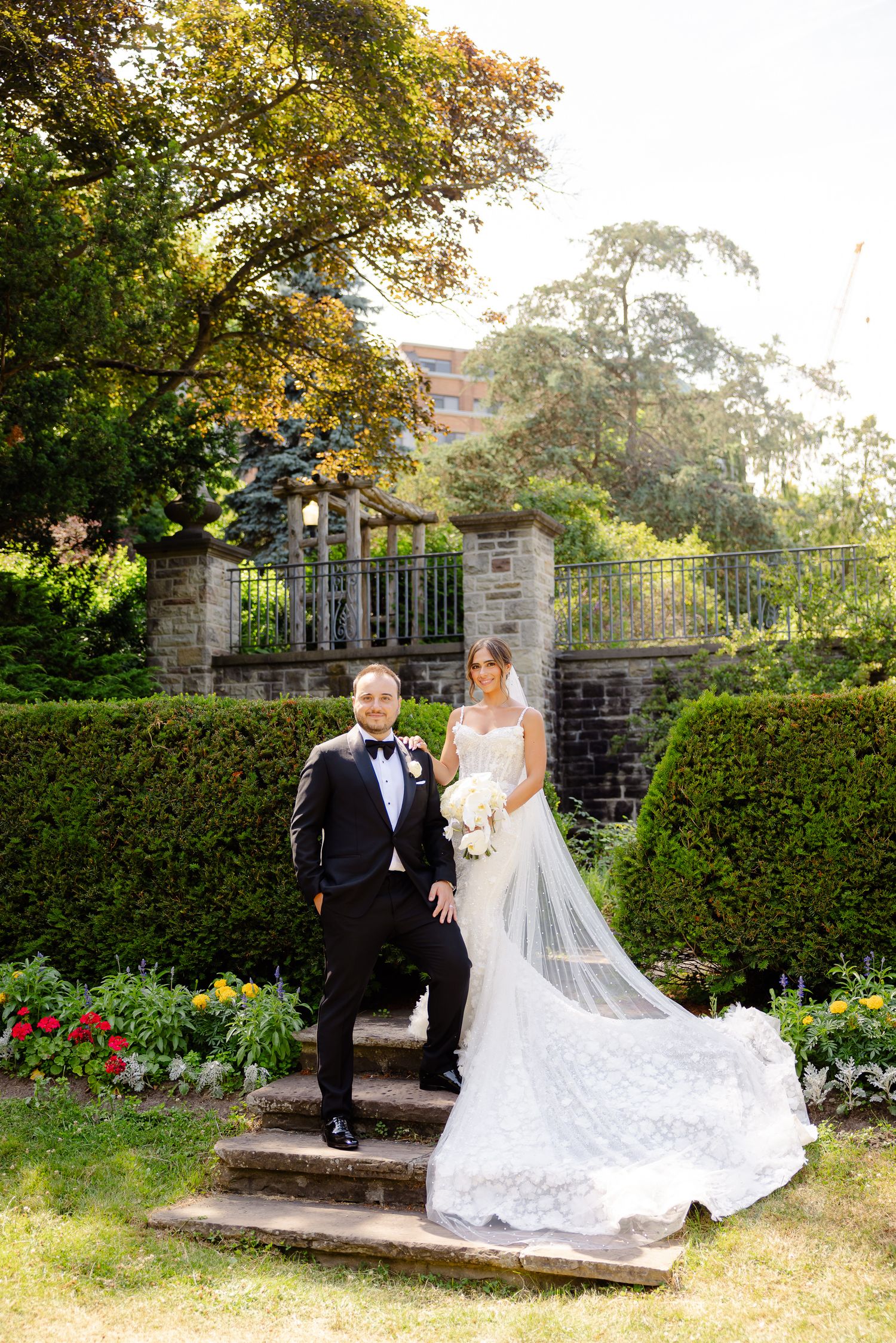 Elegant wedding couple poses on stone steps by flowering garden and ornate gate in late afternoon sunlight.