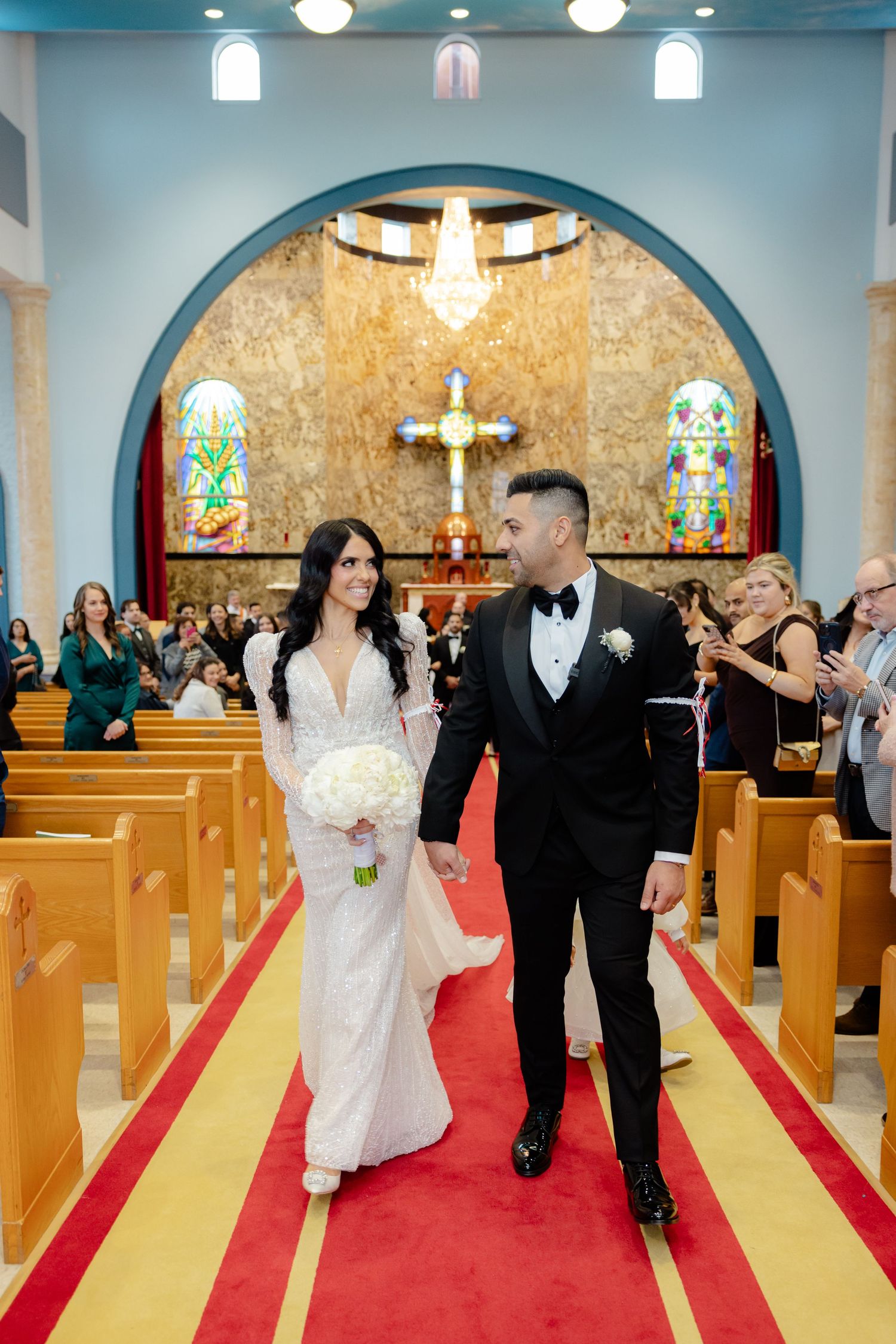Couple walks down red and gold church aisle while guests applaud during recessional.