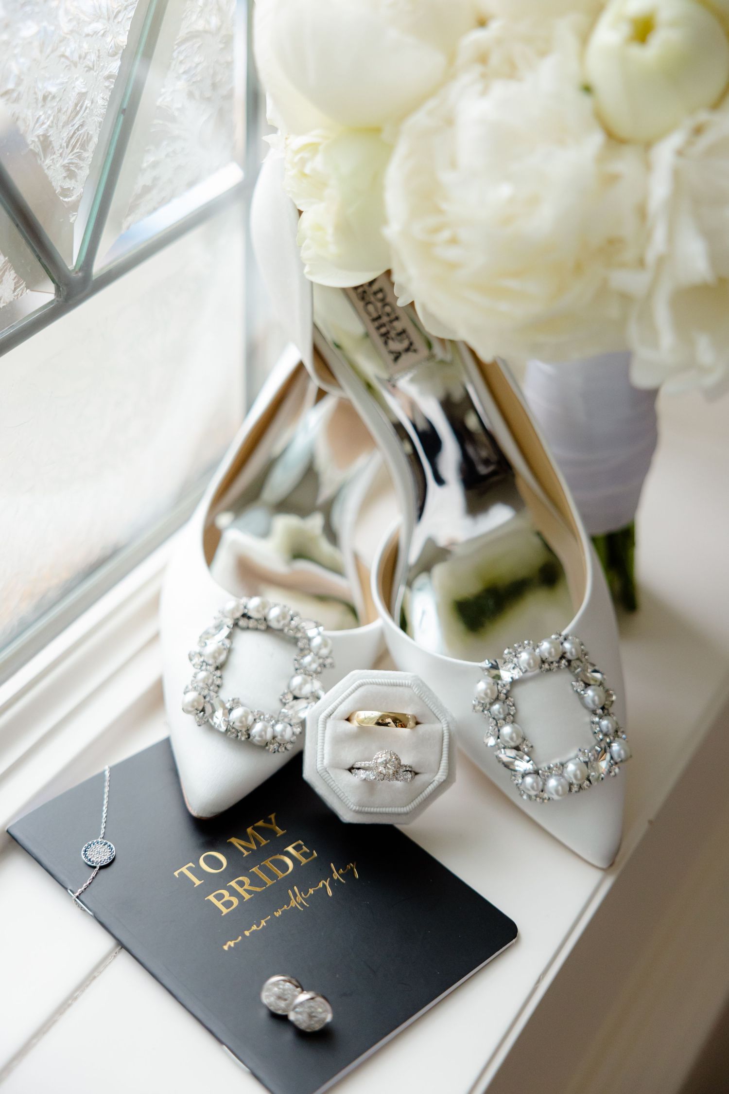 Wedding shoes, rings, and a black 'To My Bride' card arranged with white flowers.