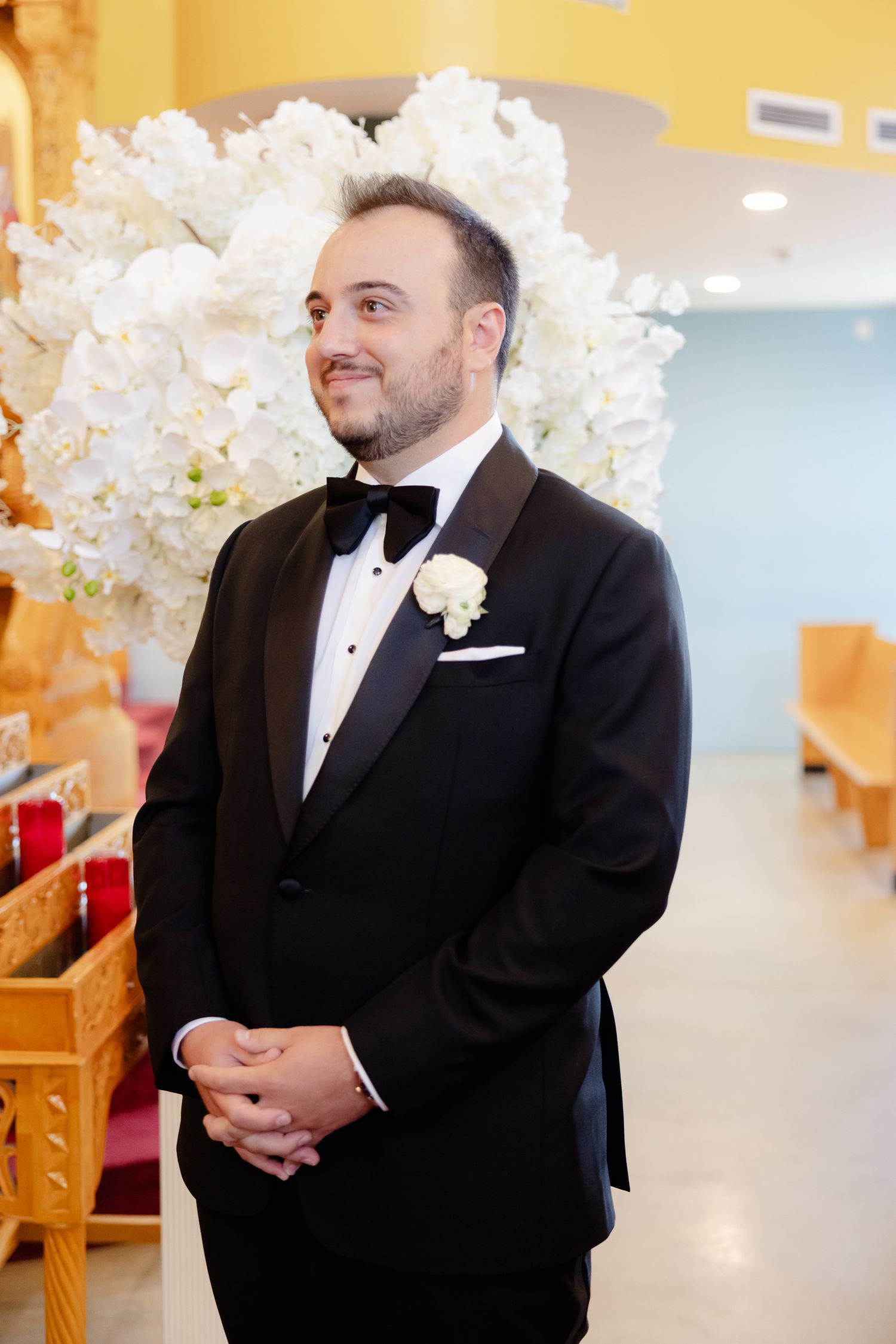 A groom wearing a classic black tuxedo and bowtie stands by white floral decorations at a wedding ceremony.