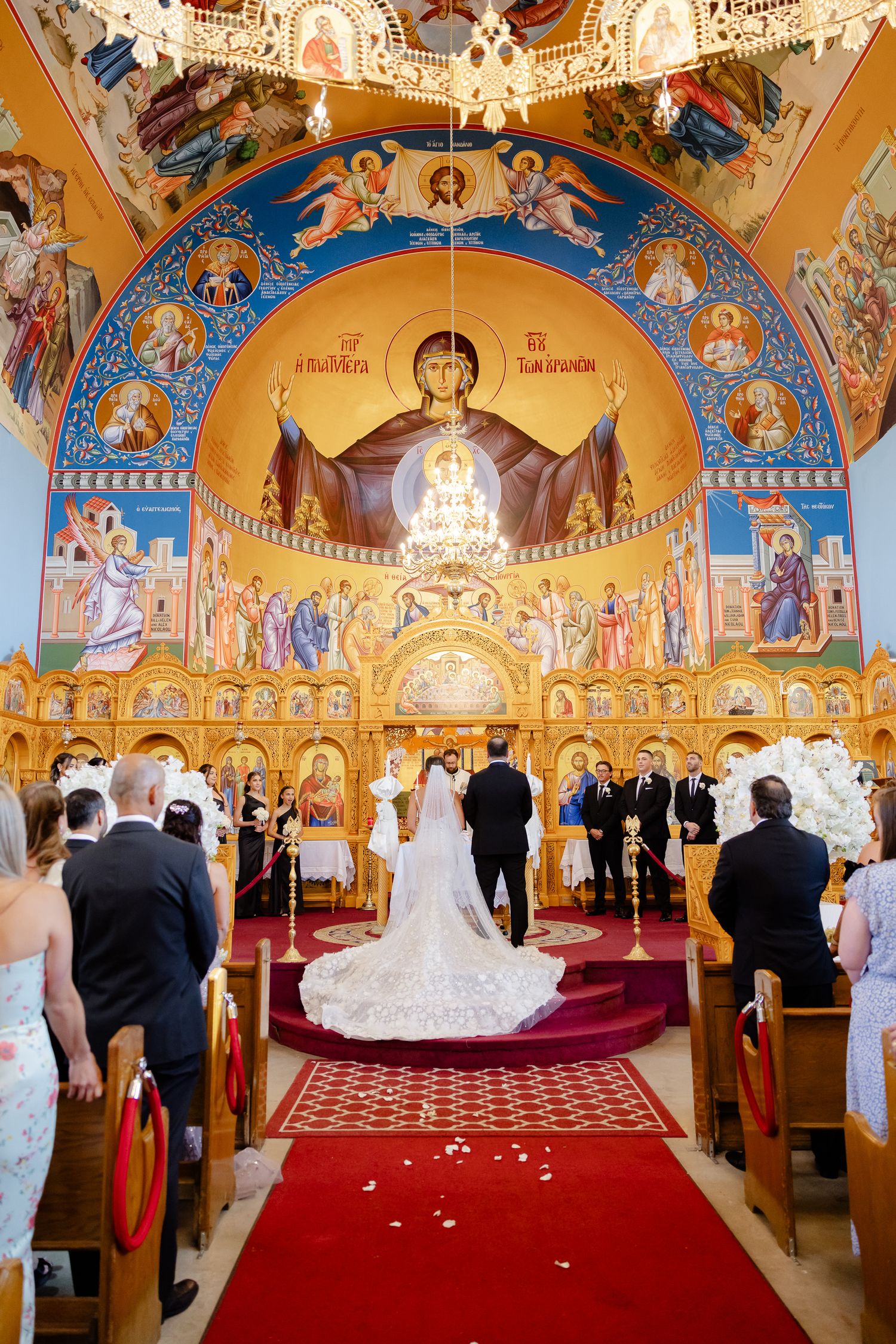 Wedding ceremony taking place in an ornate Orthodox church with golden walls and religious frescoes on a dramatic red carpet.