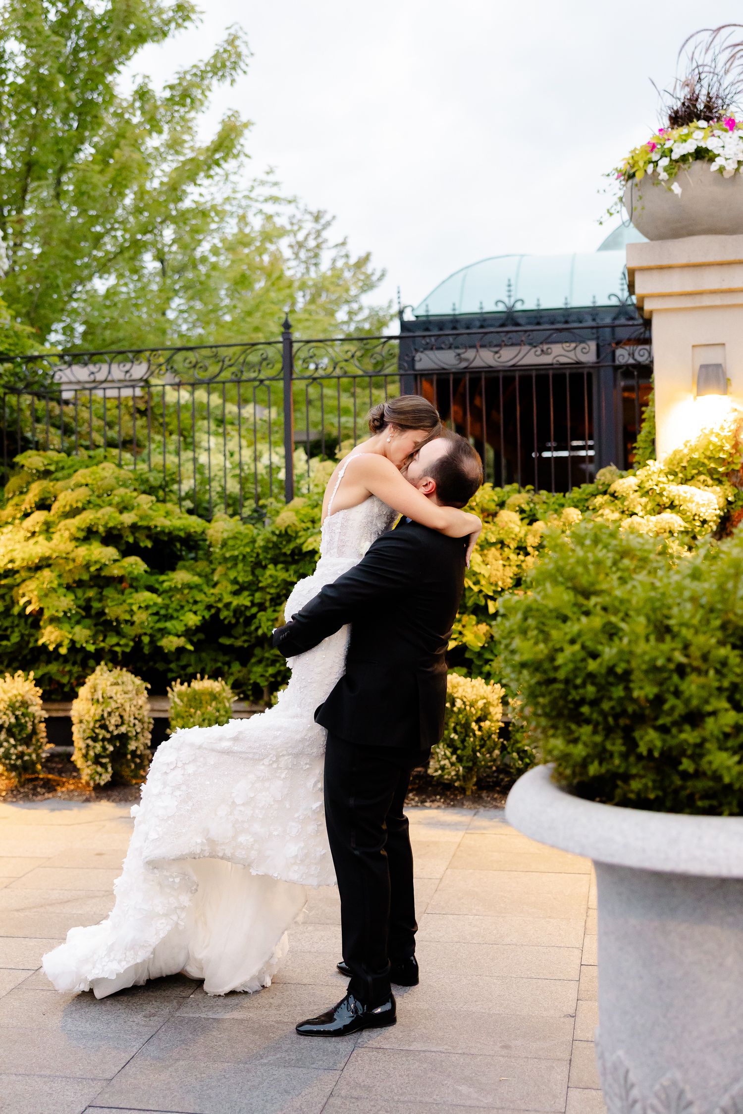 A couple sharing a romantic embrace outdoors in a garden setting with yellow foliage and decorative planters.
