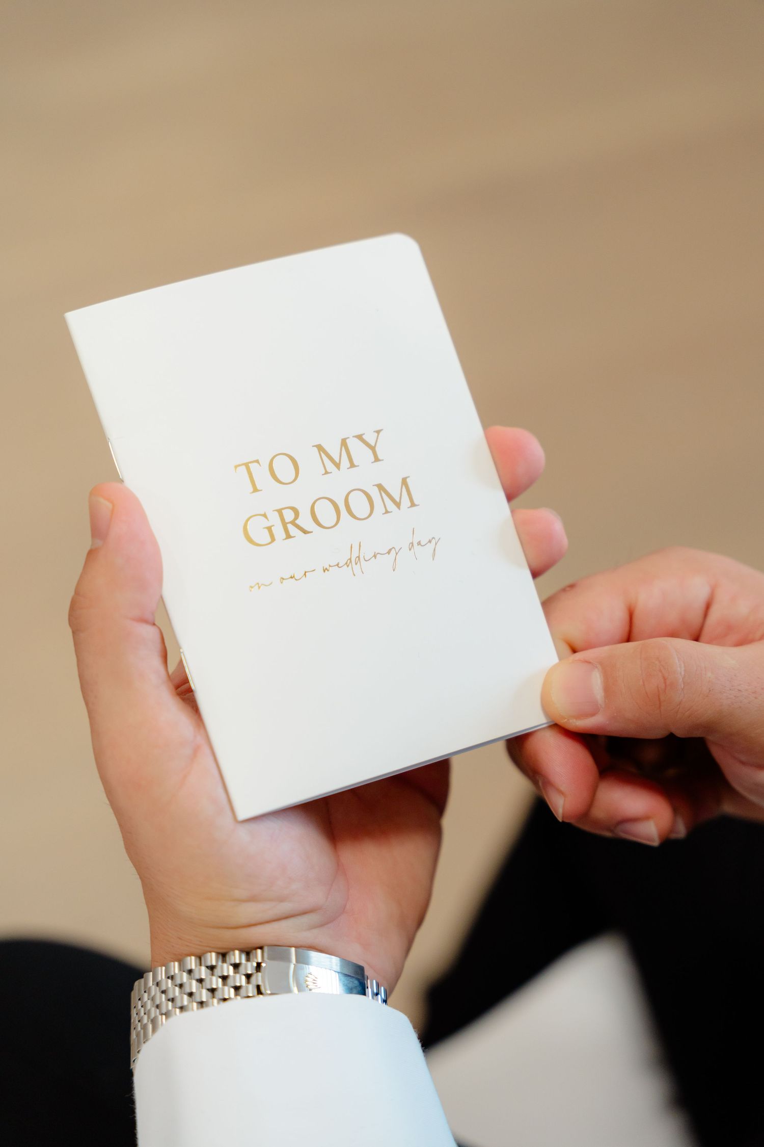 White card with gold text reading 'TO MY GROOM' held by hands wearing formal cuffs.