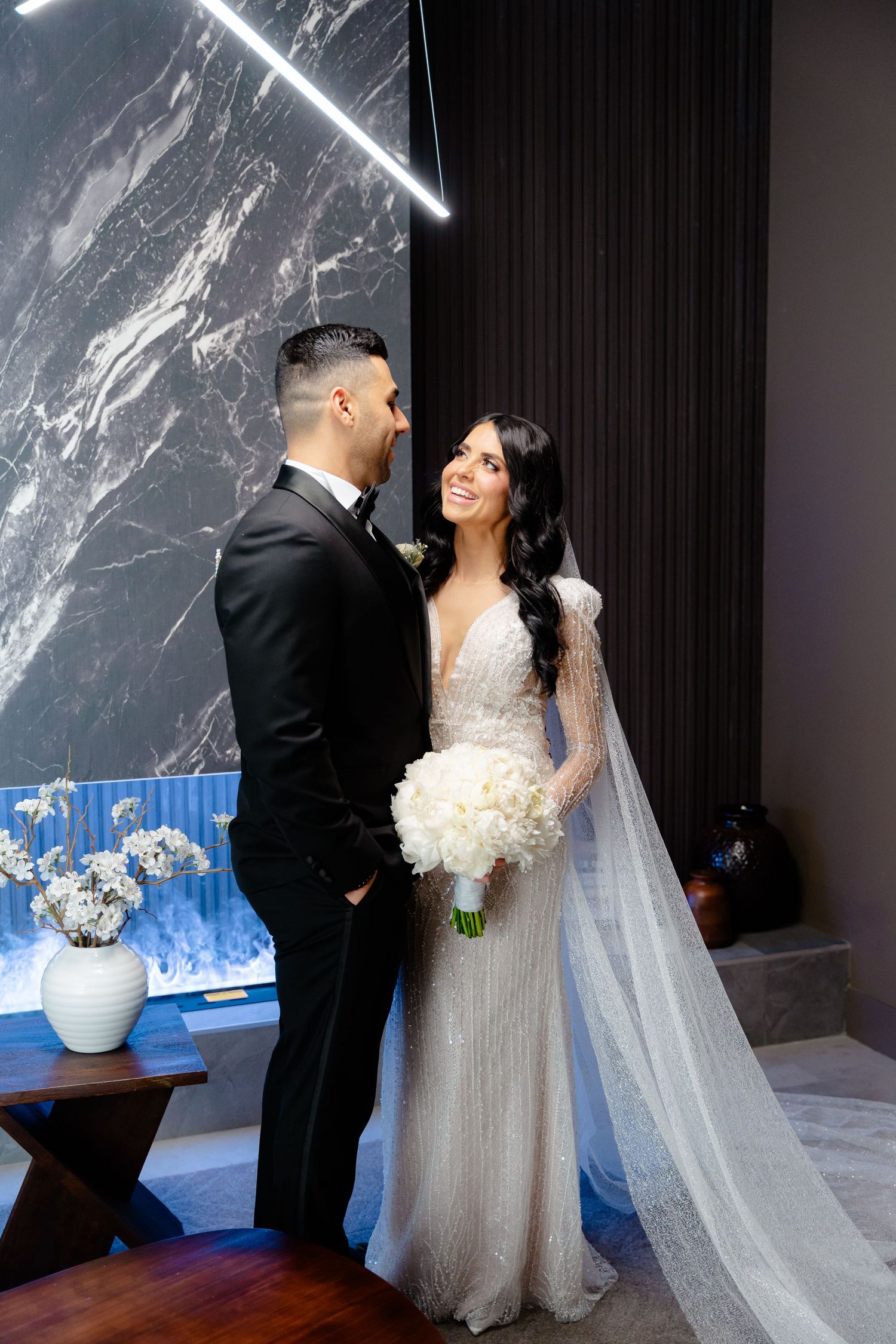 A bride and groom share a loving glance in front of a marble wall and blue decor.