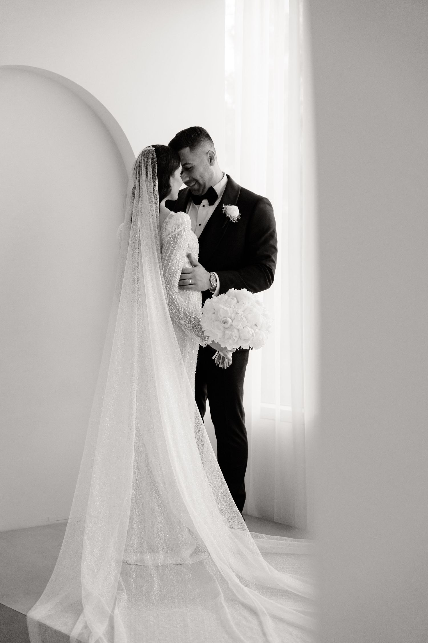 Romantic black and white portrait of newlyweds embracing by window with flowing veil.