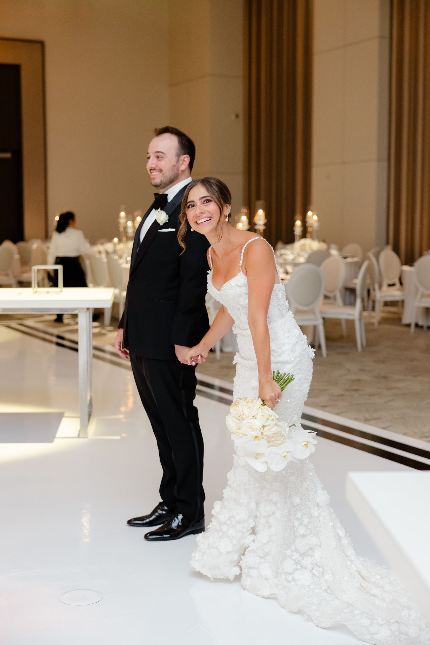 Wedding couple share playful moments in an elegant white reception space with candlelit tables and modern decor.